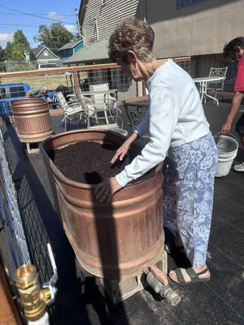 An elderly woman with short hair, wearing a white top and patterned pants, plants soil into a large terracotta pot on a patio.