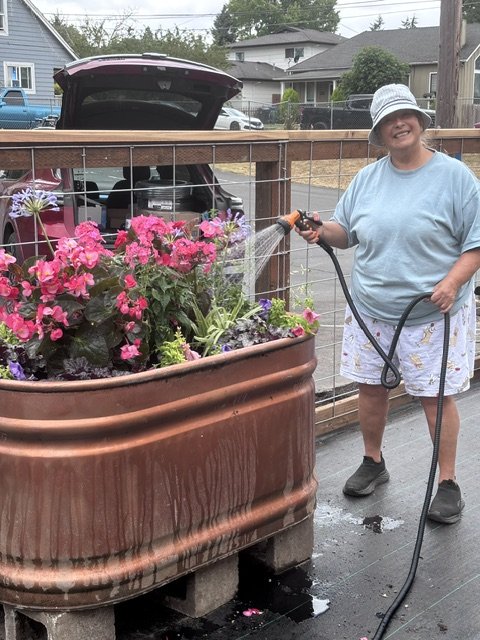 A woman watering pink and purple flowers in a large copper-colored planter on a paved outdoor surface, wearing a light blue shirt, shorts, and a bucket hat, with a residential neighborhood in the background.