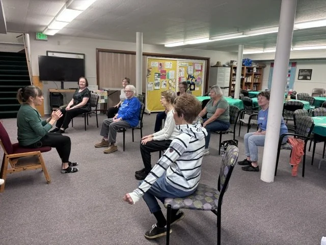 A group of people participating in a seated workshop or discussion in a community room, sitting on chairs arranged in a semi-circle.