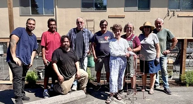 Group of ten people standing and kneeling outside in front of a building, some smiling, two women with flowers and hats, seven men in casual clothing.