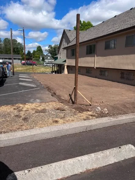 A wooden post is set in a patch of dirt next to a parking lot and sidewalk in front of a beige building with multiple windows. The sky is partly cloudy.