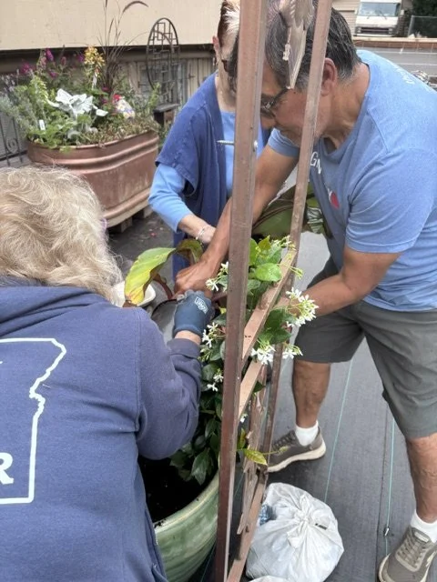 Three people working together to plant or tend flowers in a large planter on a balcony or rooftop garden.