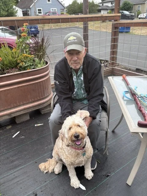 An elderly man with a baseball cap sitting on a deck with his light-colored, fluffy dog. There are potted plants, a table with objects on it, and a parking lot with houses in the background.