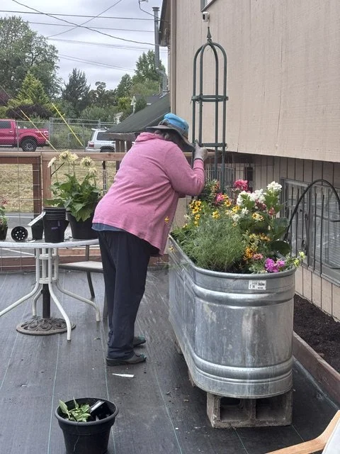 Woman in pink jacket and blue hat watering flowers in a large metal container on a porch.