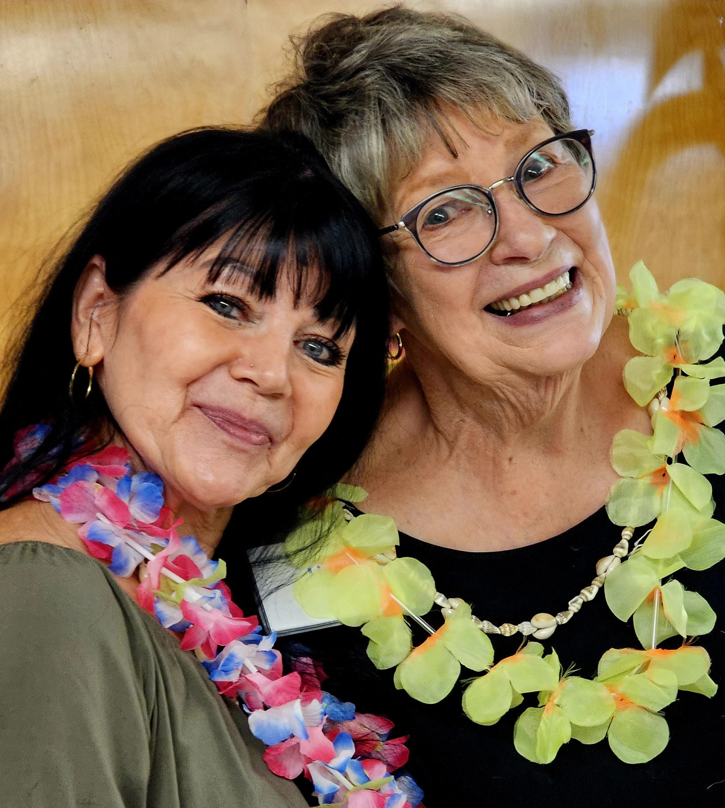Close-up of two smiling women wearing leis, one with dark hair and the other with short gray hair and glasses, in front of a wooden background.