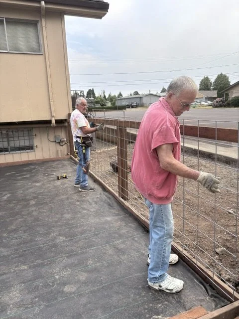 Two men working on a wire fence next to a house, on a paved surface, with a street and trees in the background.