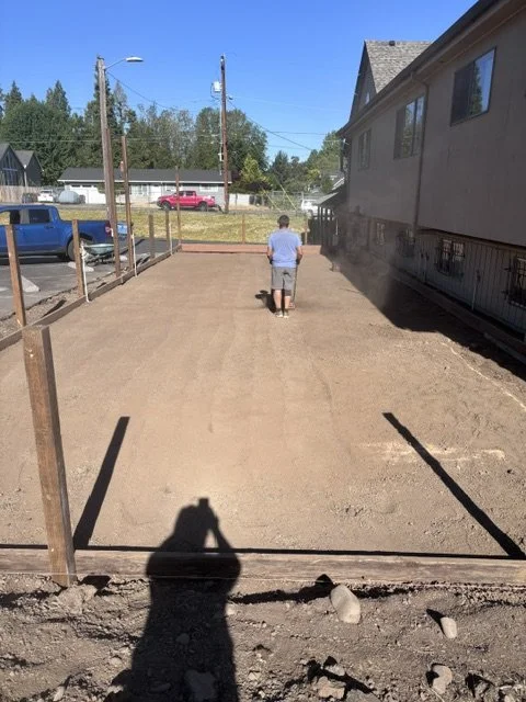 A person is leveling soil in a backyard with a hand compactor, preparing the area for paving or landscaping, with a cloud of dust rising behind them.