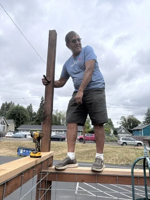 A man working on a construction project outdoors, standing on a wooden platform, holding a power drill, with a partially built wooden structure in front of him.