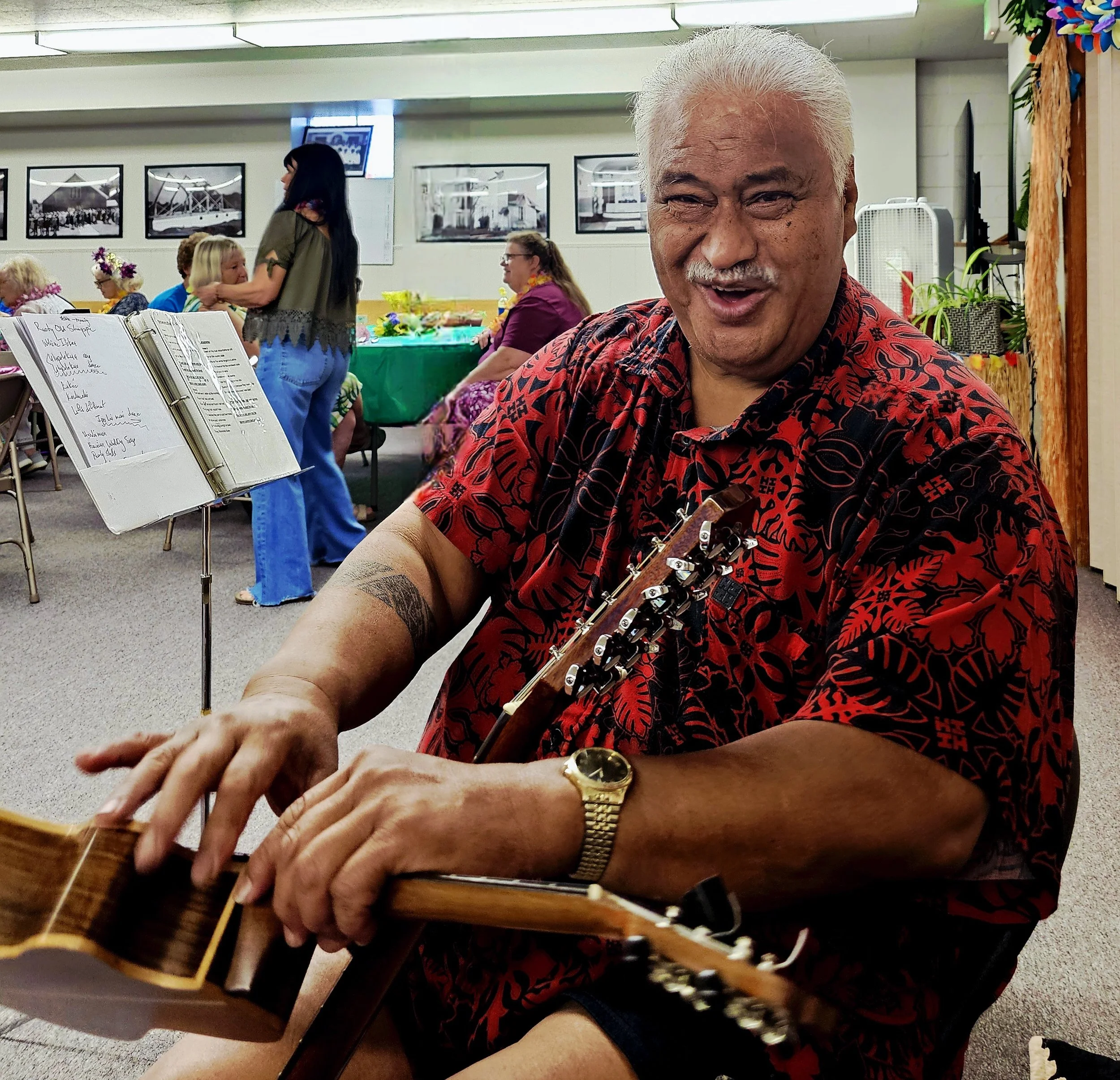 A cheerful man with gray hair and tattoos, wearing a red and black patterned shirt and gold watch, sitting at a table with a guitar in his hands at a lively indoor gathering with people seated around tables decorated with tropical flowers.