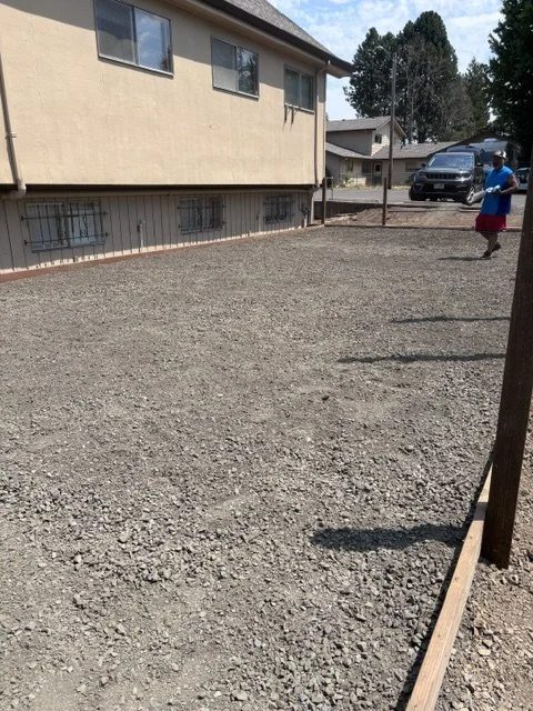 Empty gravel yard with a house in the background, a person walking, and cars parked nearby.