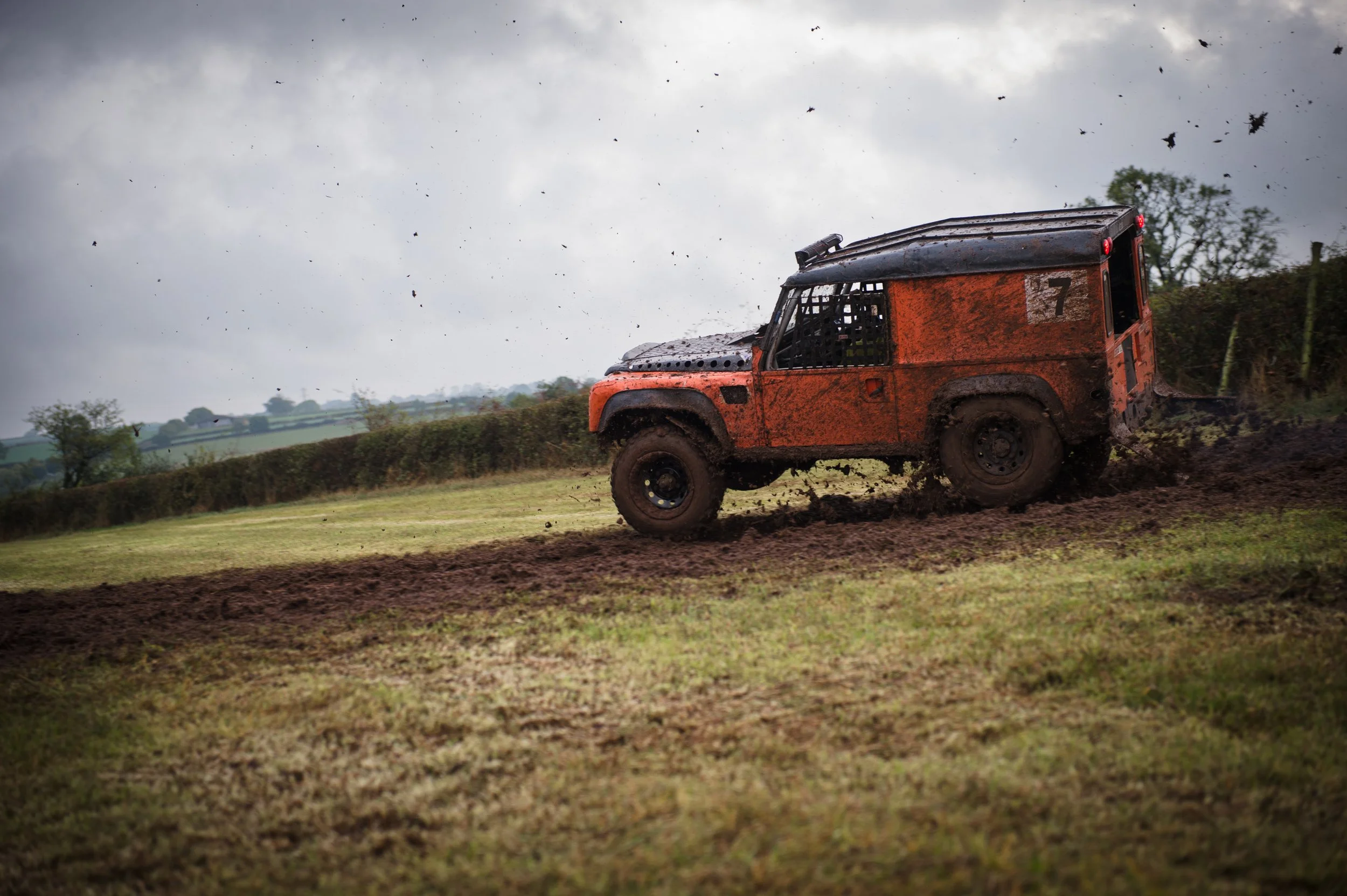 An orange off-road vehicle kicking up dirt on a muddy track, with dark clouds and trees in the background.