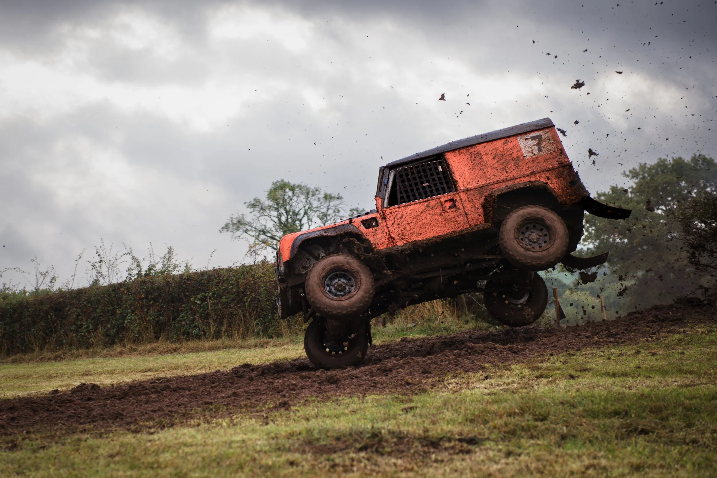 An orange off-road vehicle, possibly a dirt or rally car, airborne during a jump on a dirt track with dark clouds and trees in the background.
