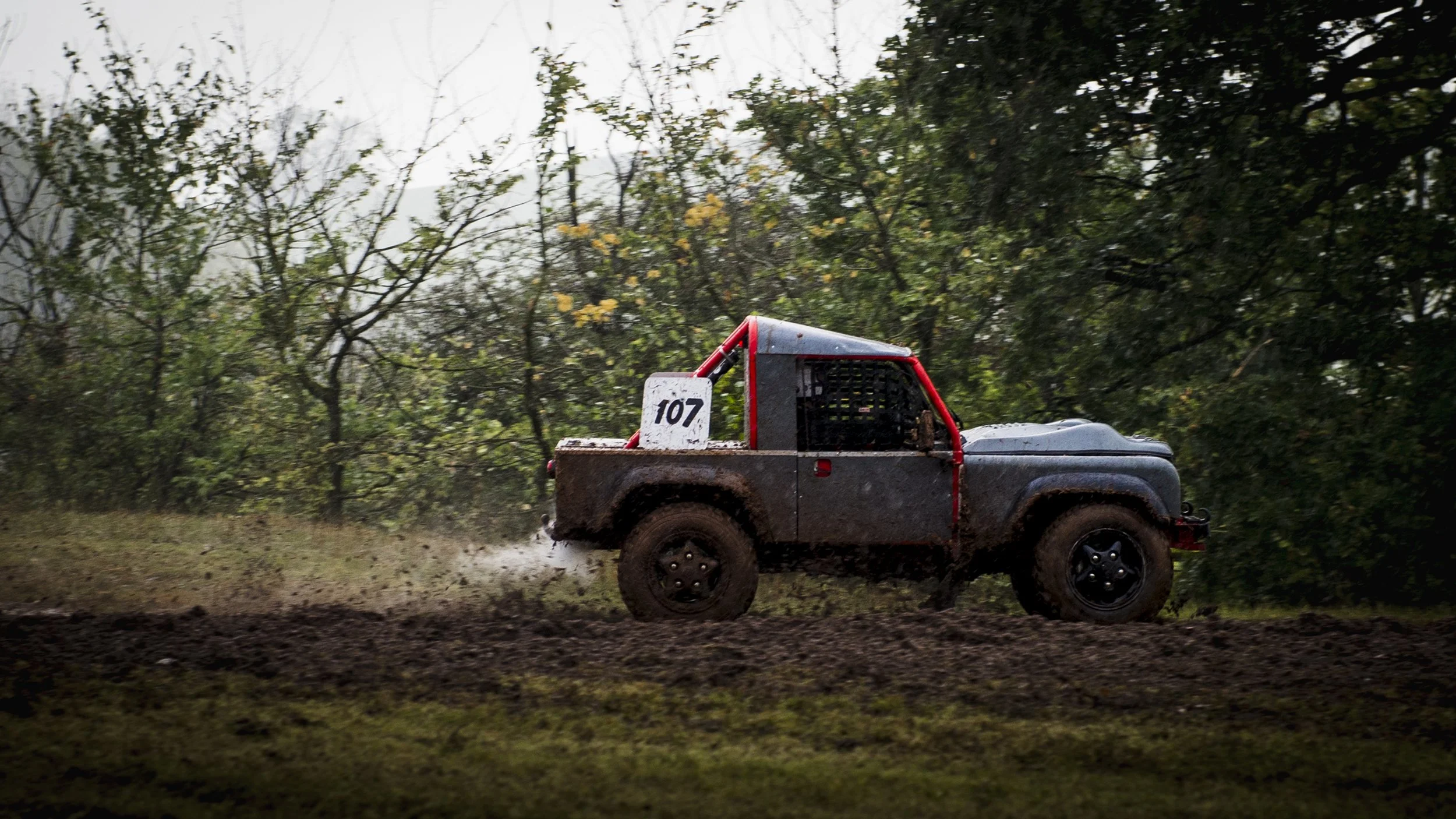 A black off-road vehicle with red accents, kicking up dirt as it drives through a muddy outdoor trail surrounded by trees and foliage.