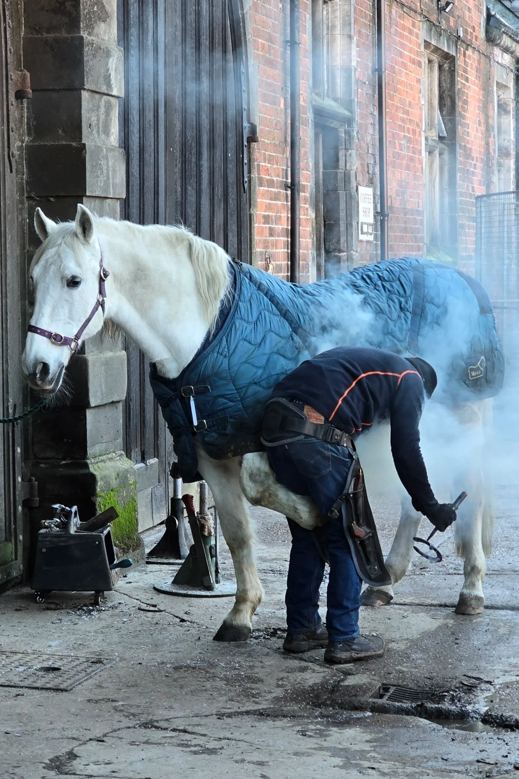 A farrier wearing a black jacket and jeans is fitting horseshoes to a white horse with a blue blanket in a courtyard next to a brick building. The horse is tied to a wall. Mist or steam is visible in the air.