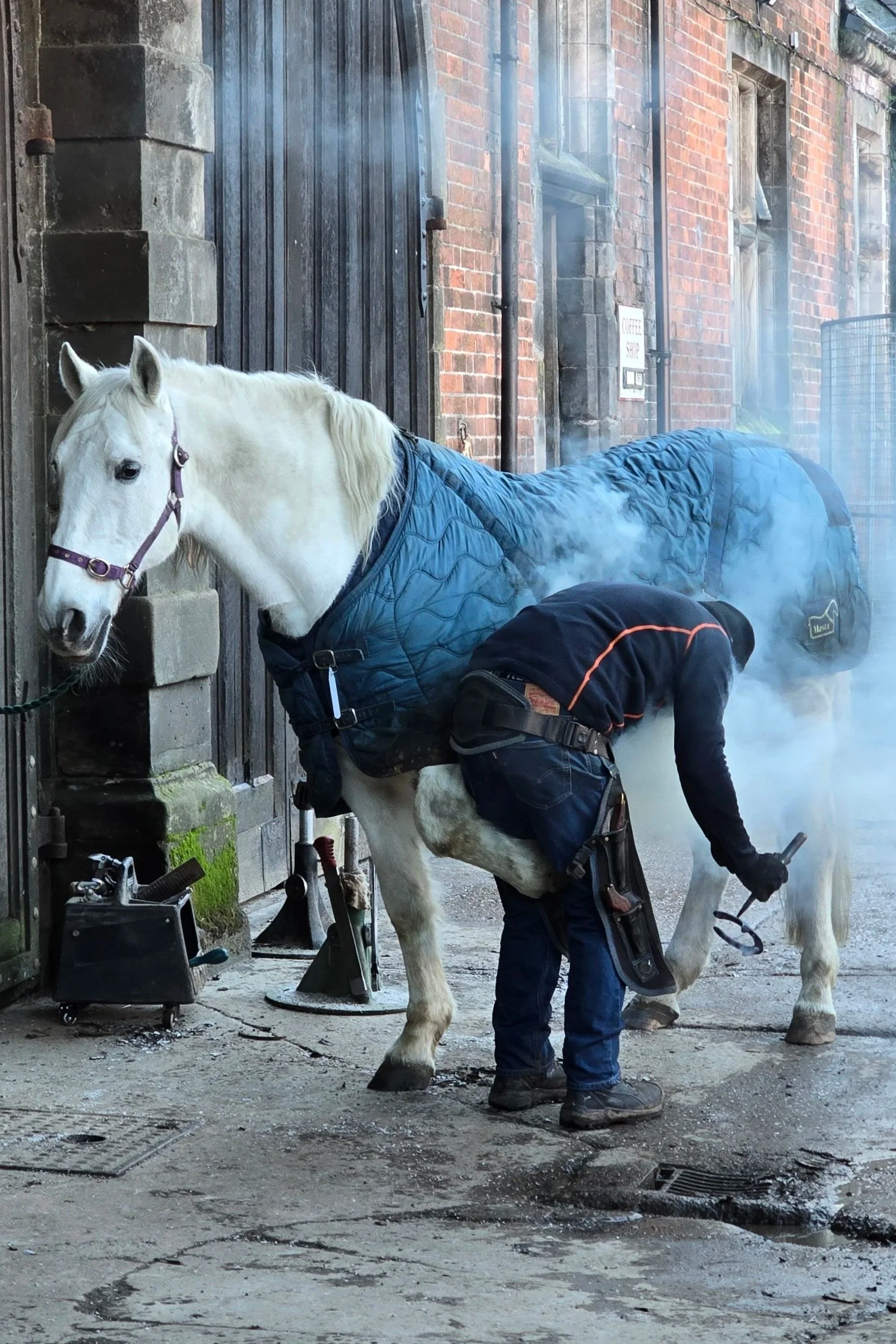 A farrier fitting horseshoes to a white horse l, which is wearing a blue blanket, outside a brick building. The horse is tied to the wall, and there is steam or smoke around them.