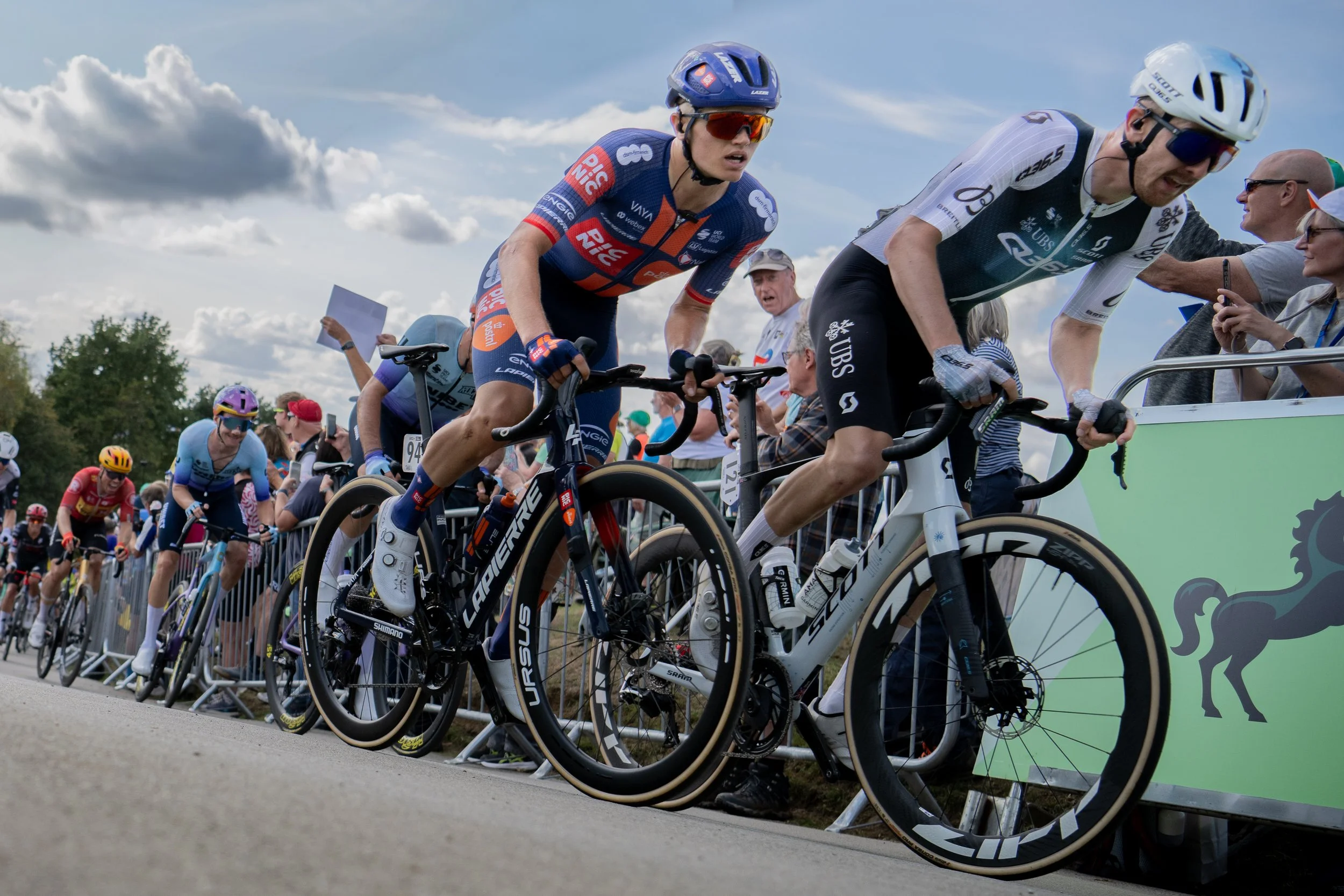Cyclists in a race update from spectators, with some riders in colorful jerseys and helmets, pushing their bikes while standing behind a barricade on a sunny day with clouds in the sky.