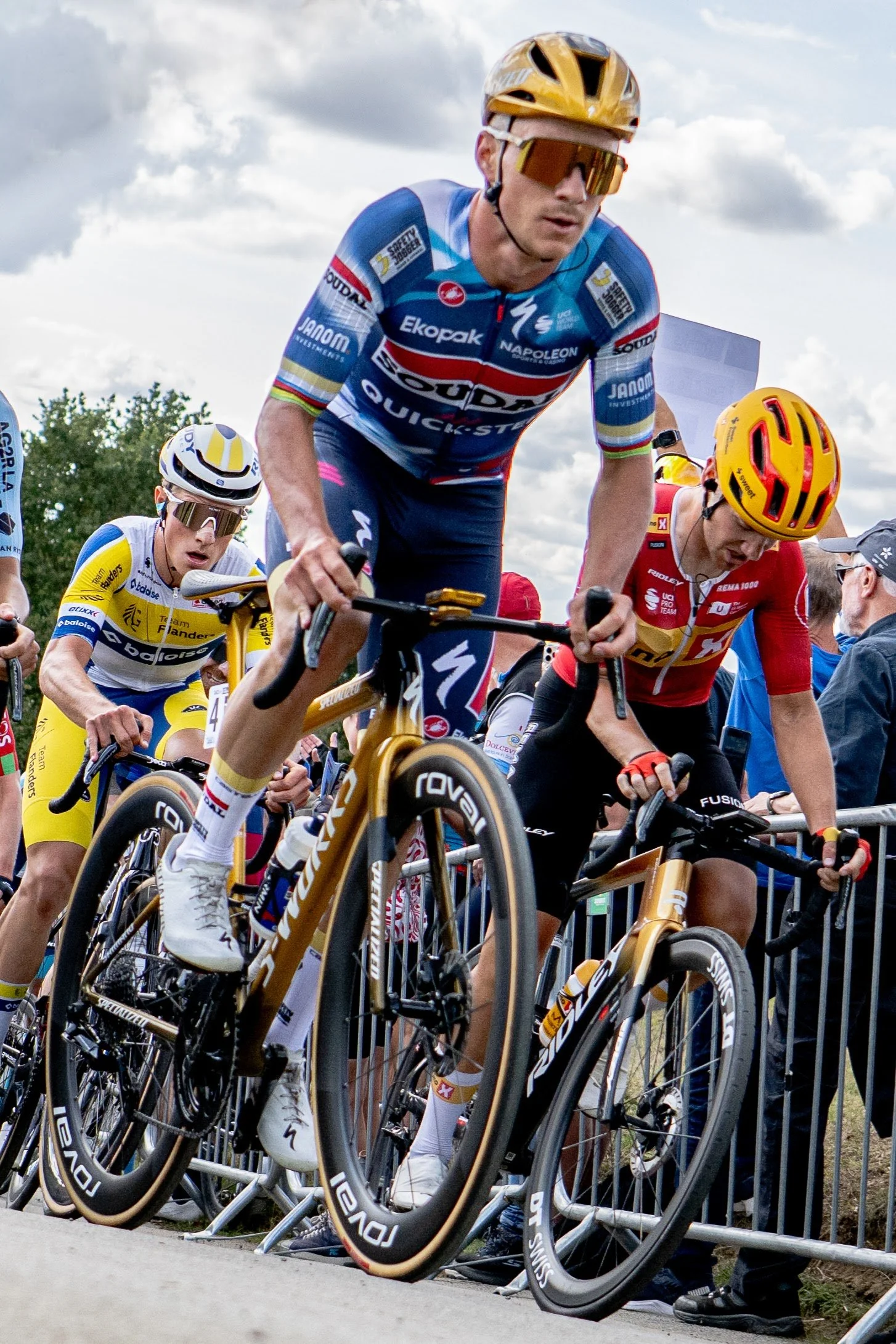 Cyclists in colorful gear preparing for a race behind start barriers, with spectators and a cloudy sky in the background.