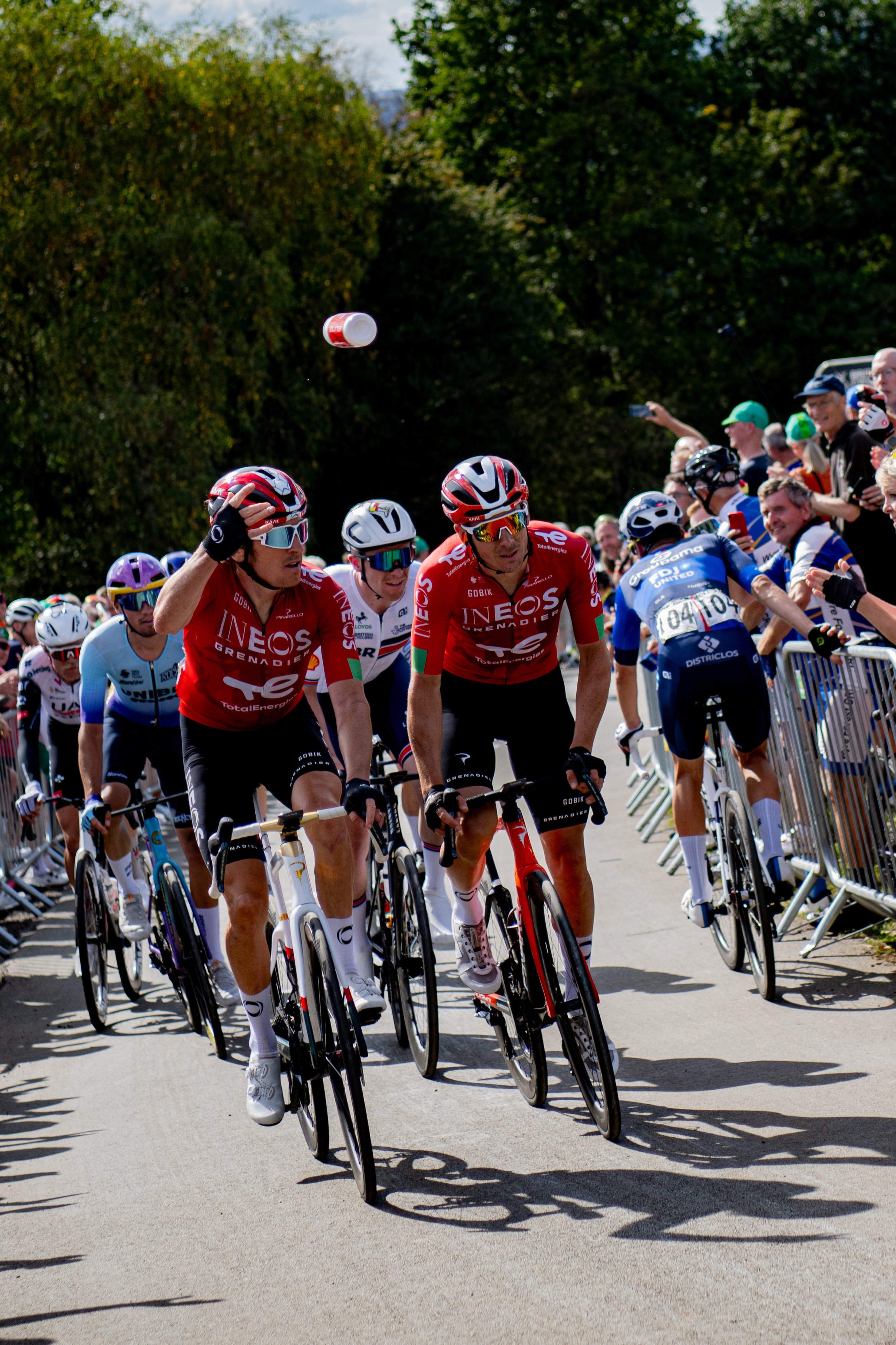 Cyclists participating in a race, wearing helmets and colorful jerseys, riding on a road with spectators on the side, some taking photos.