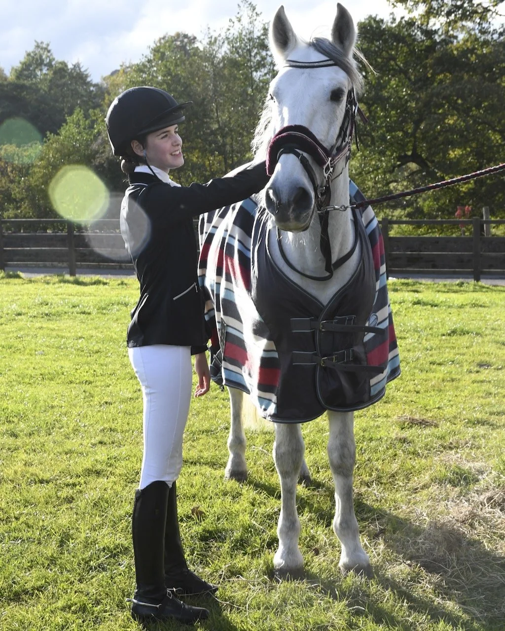 A young girl in riding attire, wearing a helmet, stands next to a white horse with a striped blanket, on a grassy field with trees and a fence in the background.