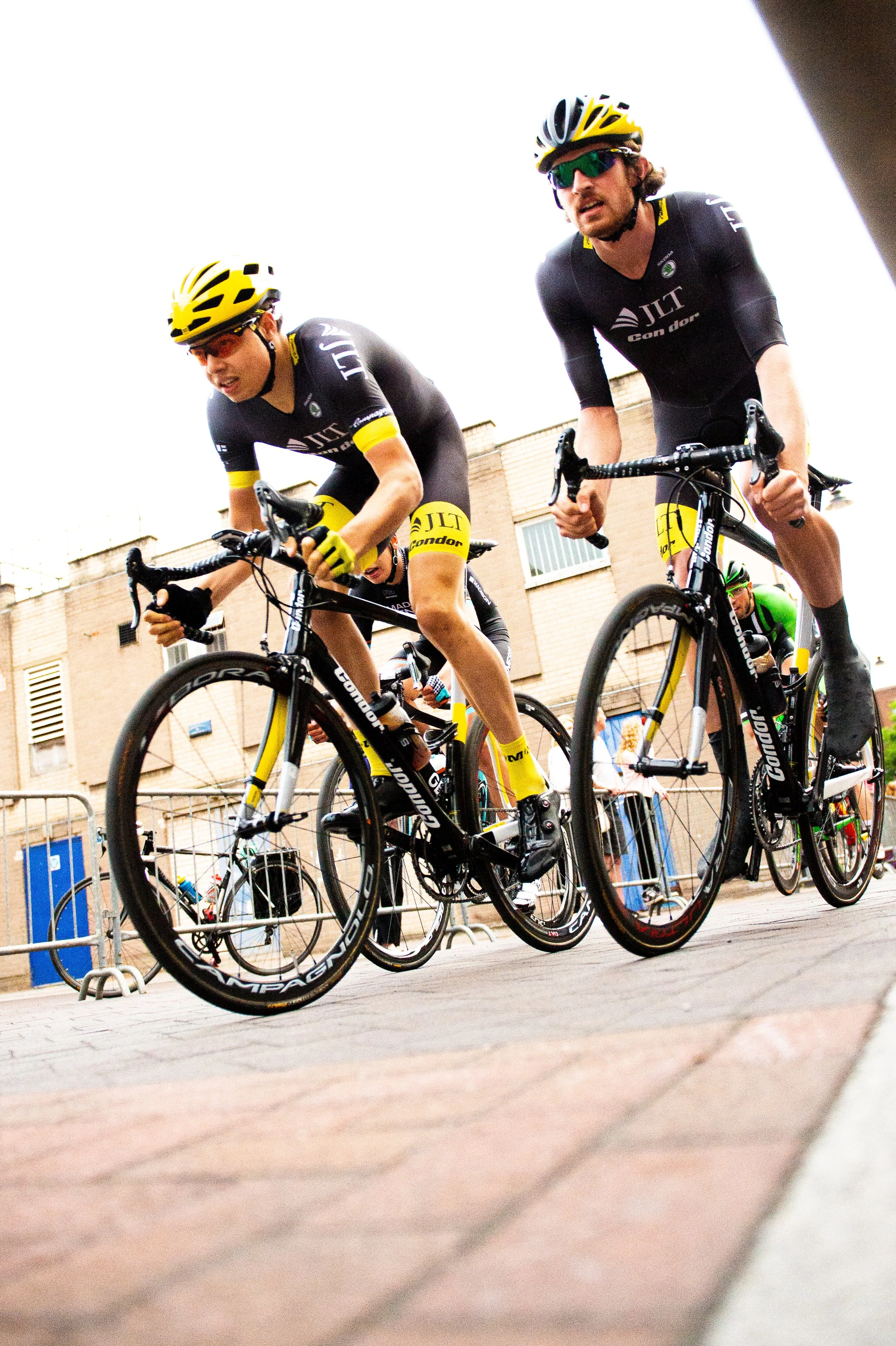 Two cyclists wearing black and yellow uniforms and helmets ride closely together on a city street. They are focused on the race, with urban buildings visible in the background.