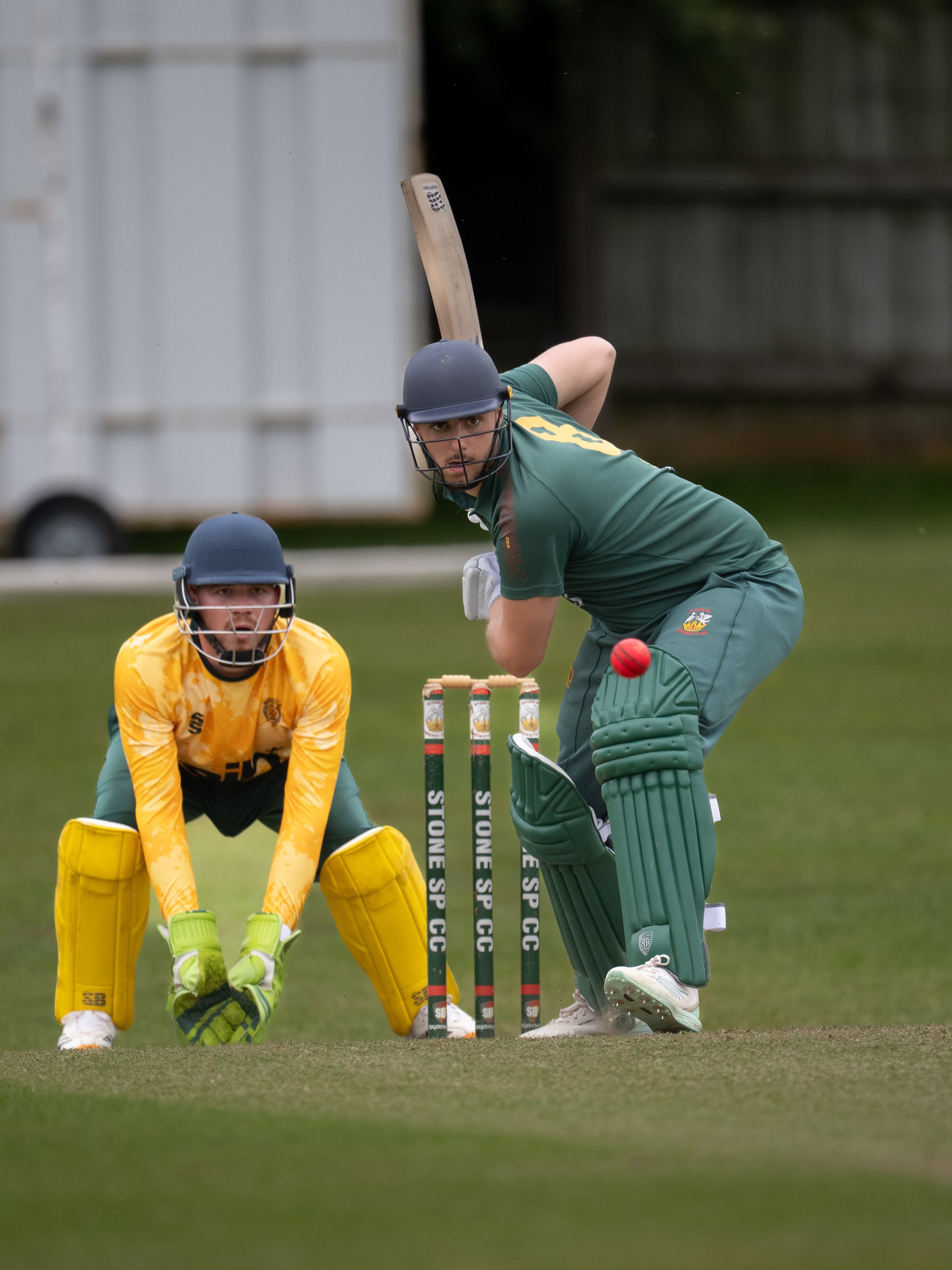 A cricket player in green uniform and helmet preparing to hit a cricket ball with a bat, while a wicketkeeper in yellow gear crouches behind the stumps on a grassy field.