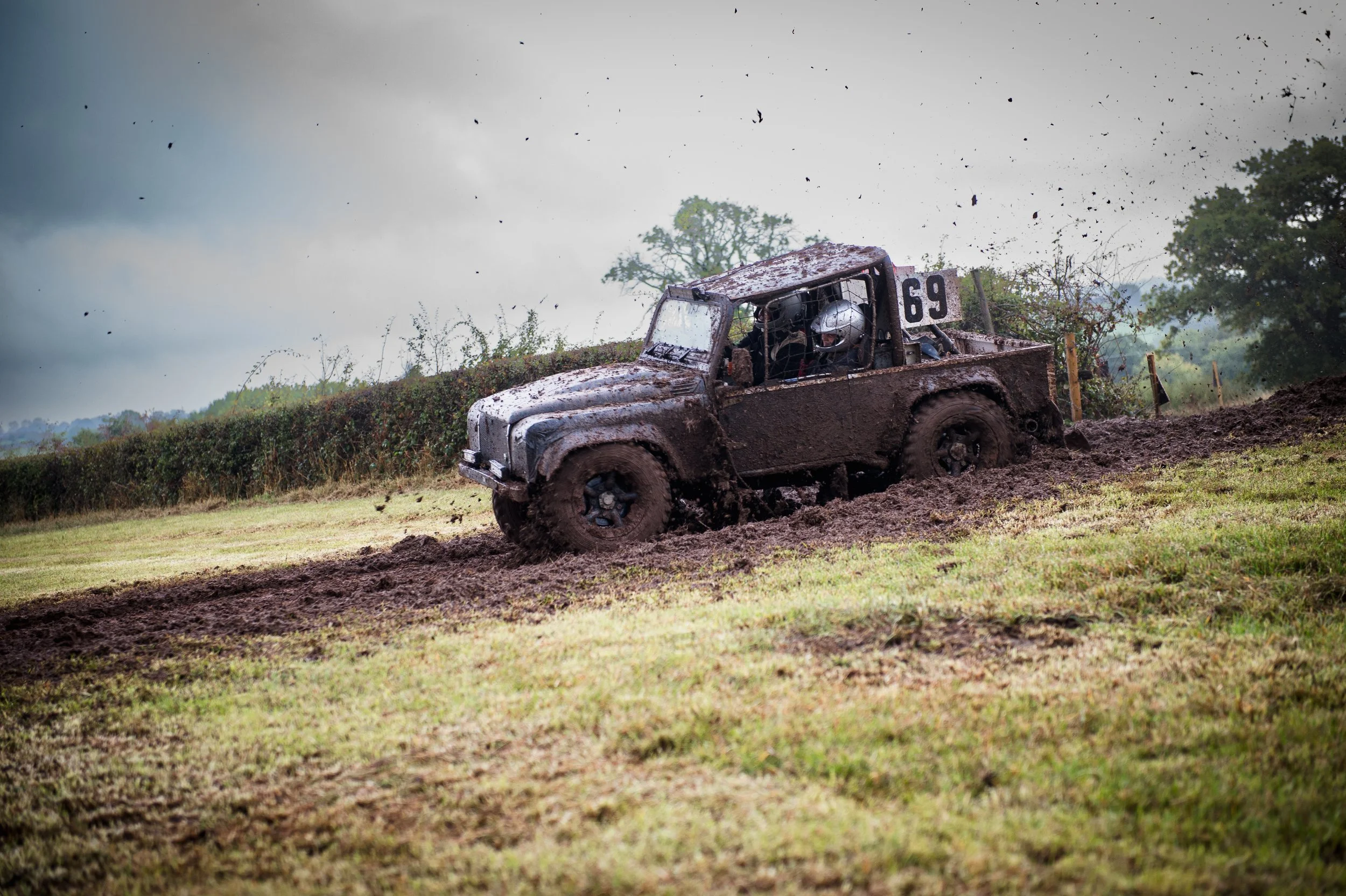 Off-road vehicle, muddy and covered in dirt, driving through a muddy terrain with a cloudy sky and trees in the background.