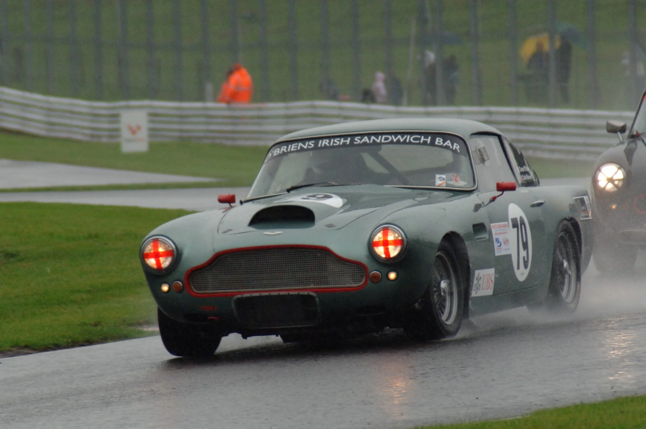 A vintage racing car with a number 15 on its side driving on a wet race track during a rainy day, with spectators and race officials in the background.