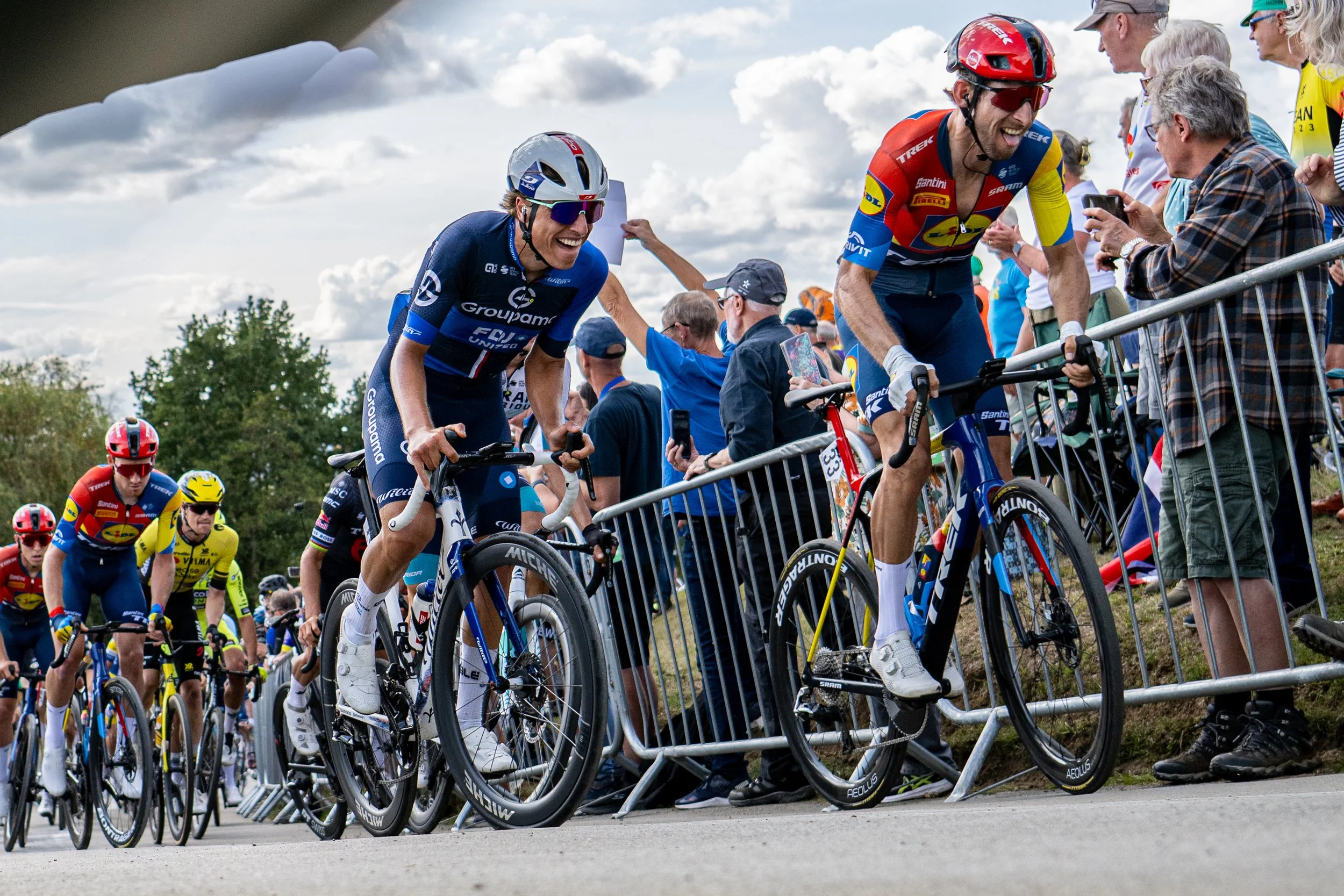 Cyclists participating in a race, with two riders at the front smiling and interacting with spectators behind a metal barrier, under a partly cloudy sky