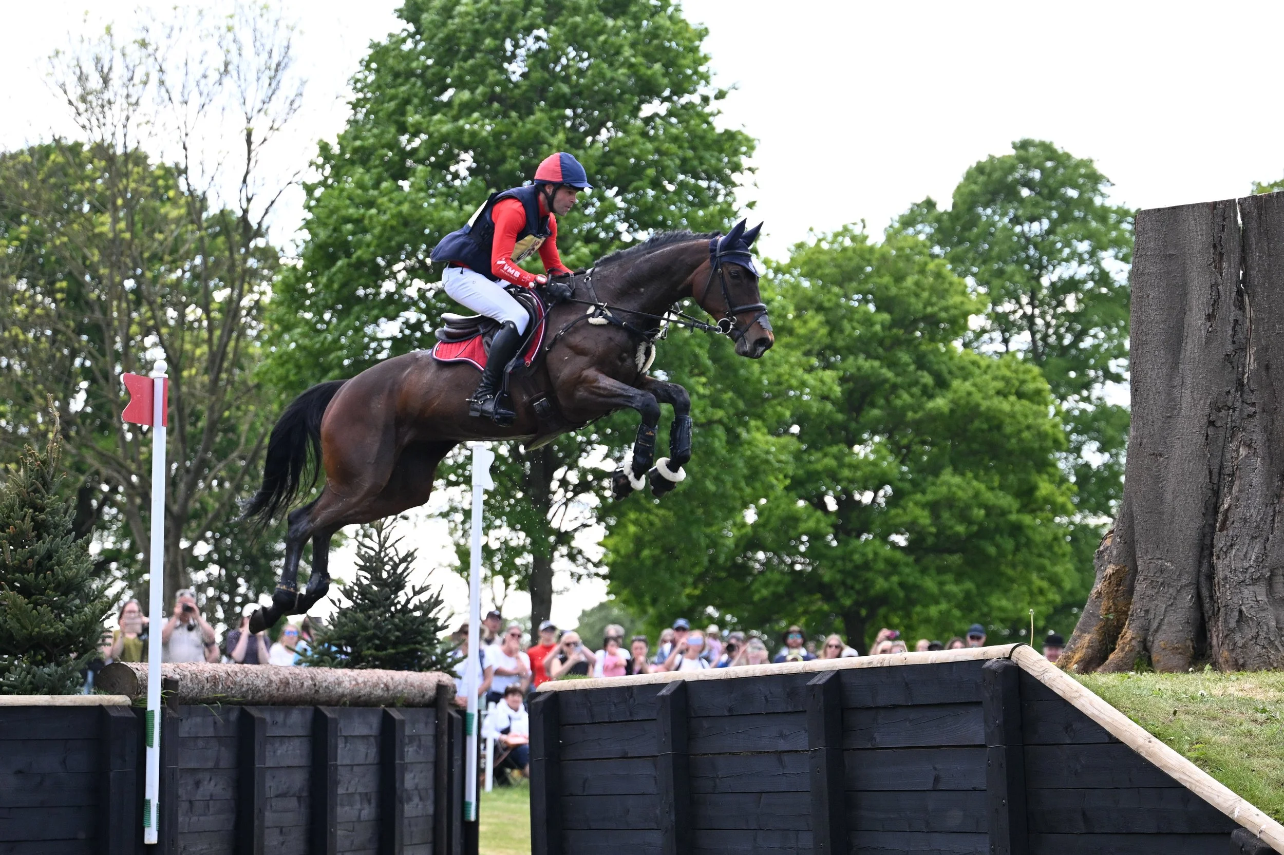 A person riding a horse during a cross-country equestrian event, jumping over a wooden obstacle with a crowd watching in the background.