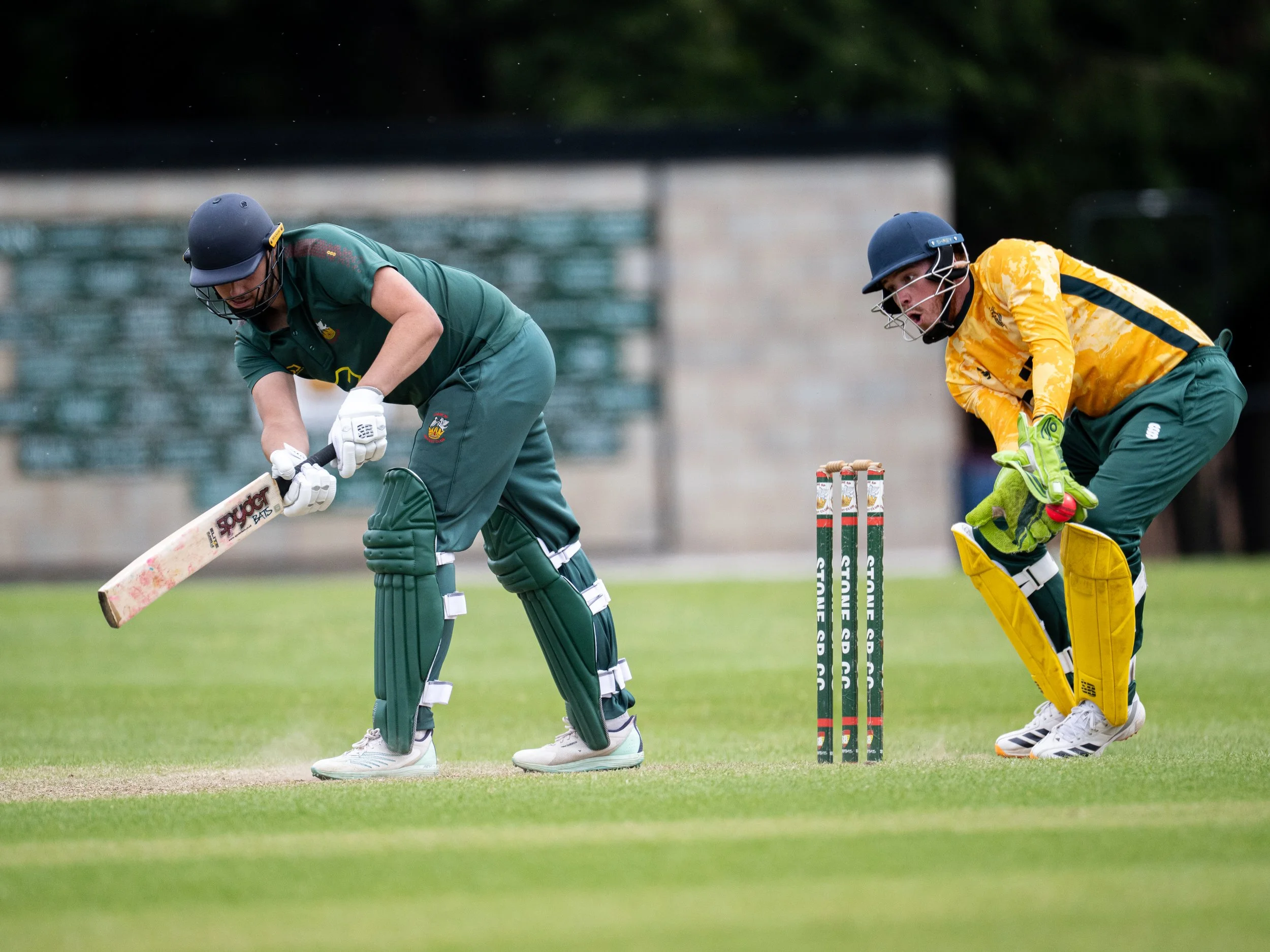 Two cricket players, one in green and the other in yellow, are on the field during a game. They are both wearing helmets and bats, preparing for the next move. The player in green is focused on the ball while the player in yellow is ready to react.