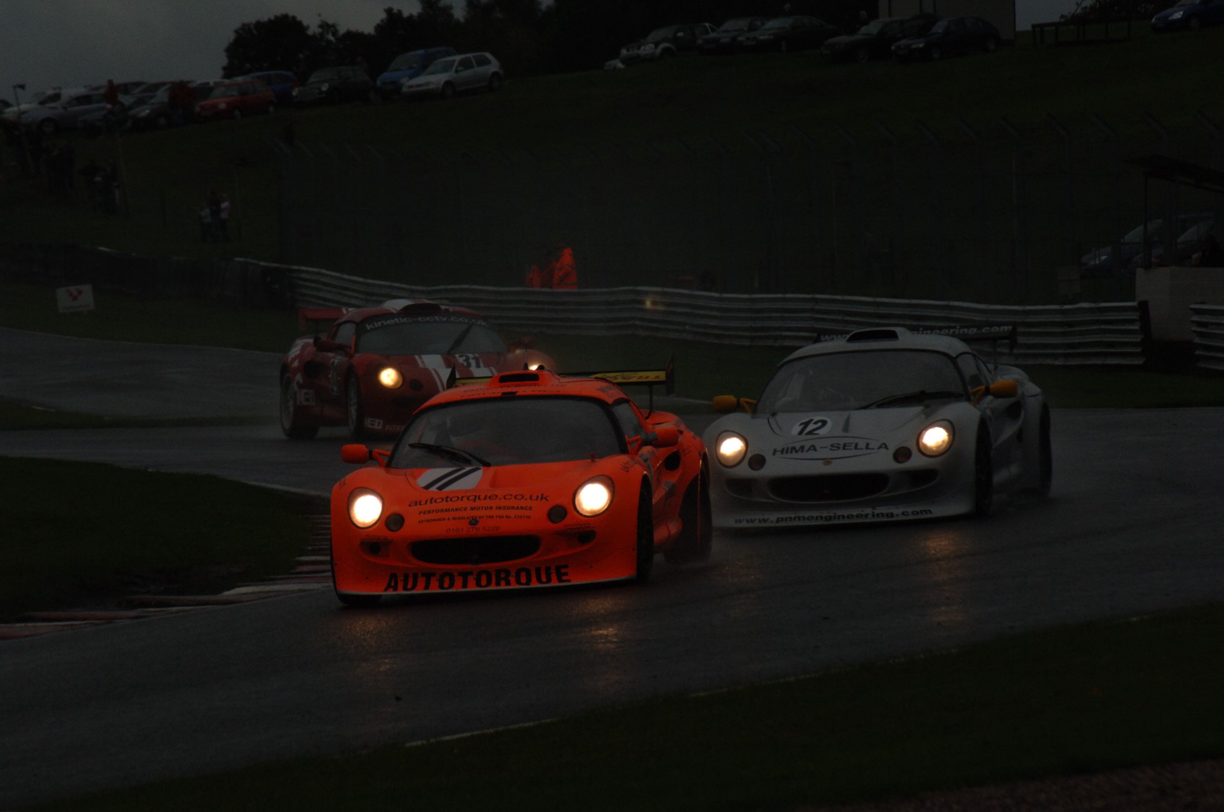 Race cars on a track during rainy weather at night, illuminated by headlights, with a background of parked cars on a hill and spectators watching.