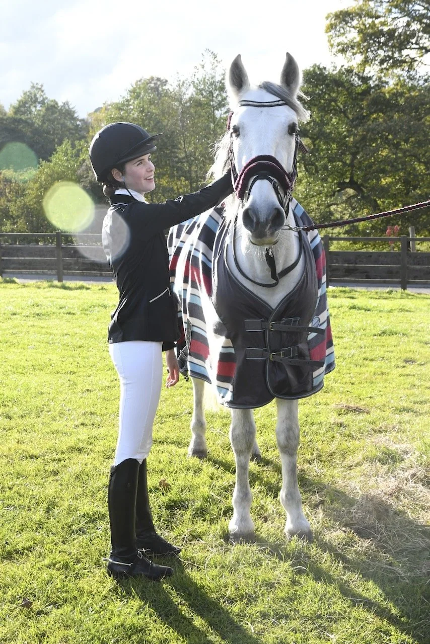 A young girl in equestrian attire, including a helmet, jacket, and riding boots, stands next to a white horse with a striped blanket and a halter, on a grassy field with trees and a wooden fence in the background.