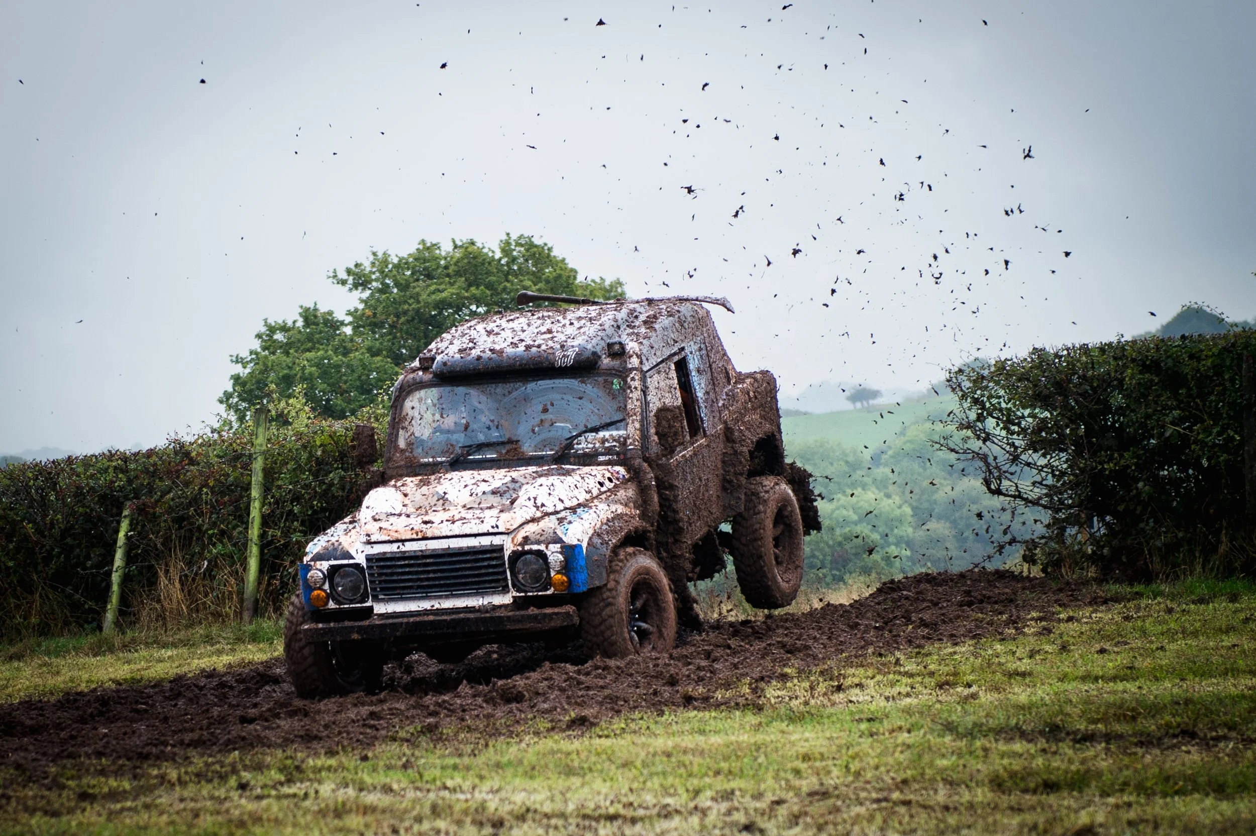 A muddy off-road Landrover vehicle, driving through a rugged dirt path surrounded by hedges and overcast sky, with mud flying overhead.