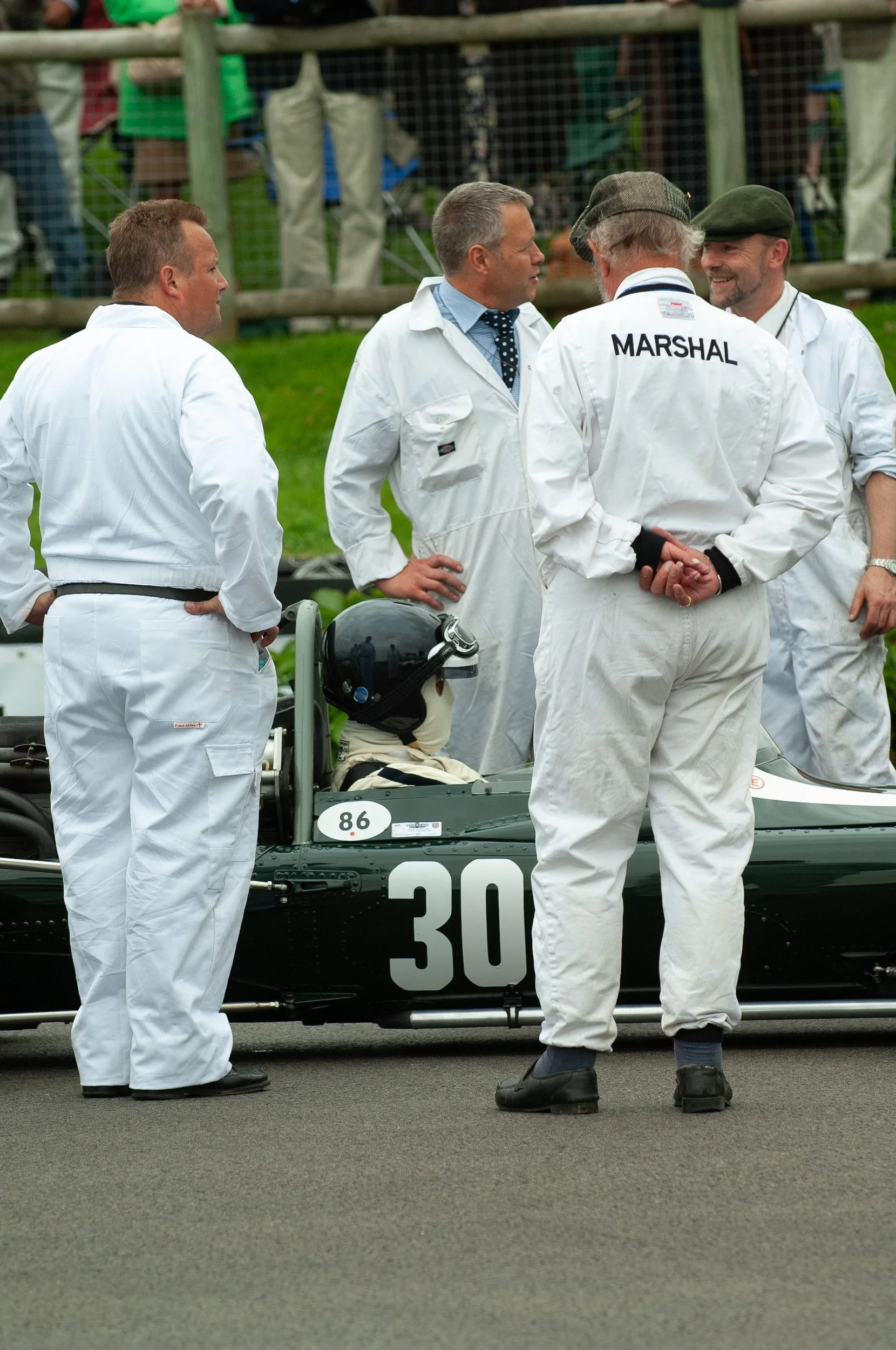 Group of men in vintage racing uniforms and hats gathered around a race car. One man wears a shirt with 'MARSHAL' on the back.