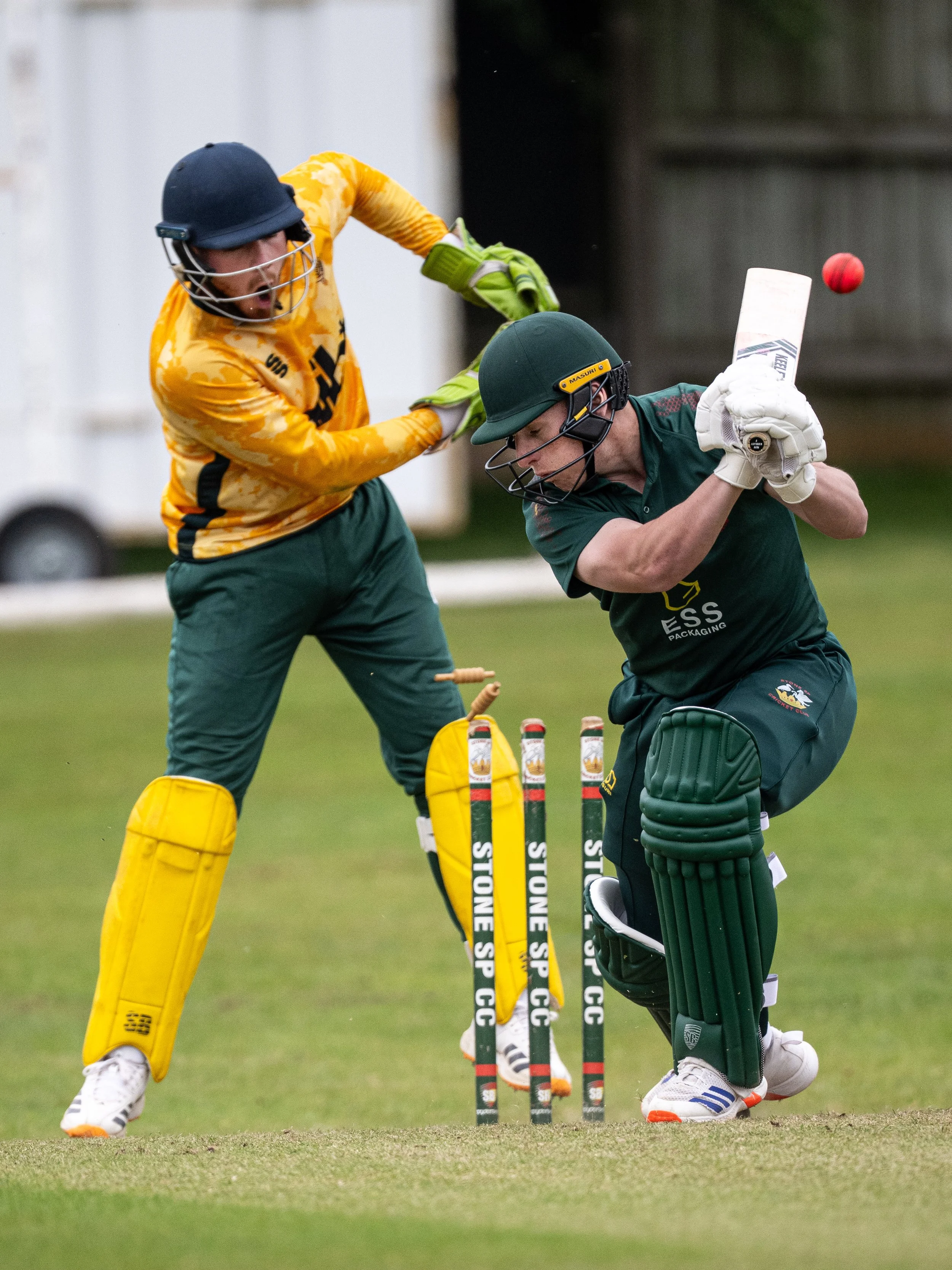 Two cricket players in protective gear and uniforms are engaged in a play during a game. One player is attempting to hit the ball with a bat, while the other is preparing to field the ball. The scene takes place on a cricket field with the stumps and