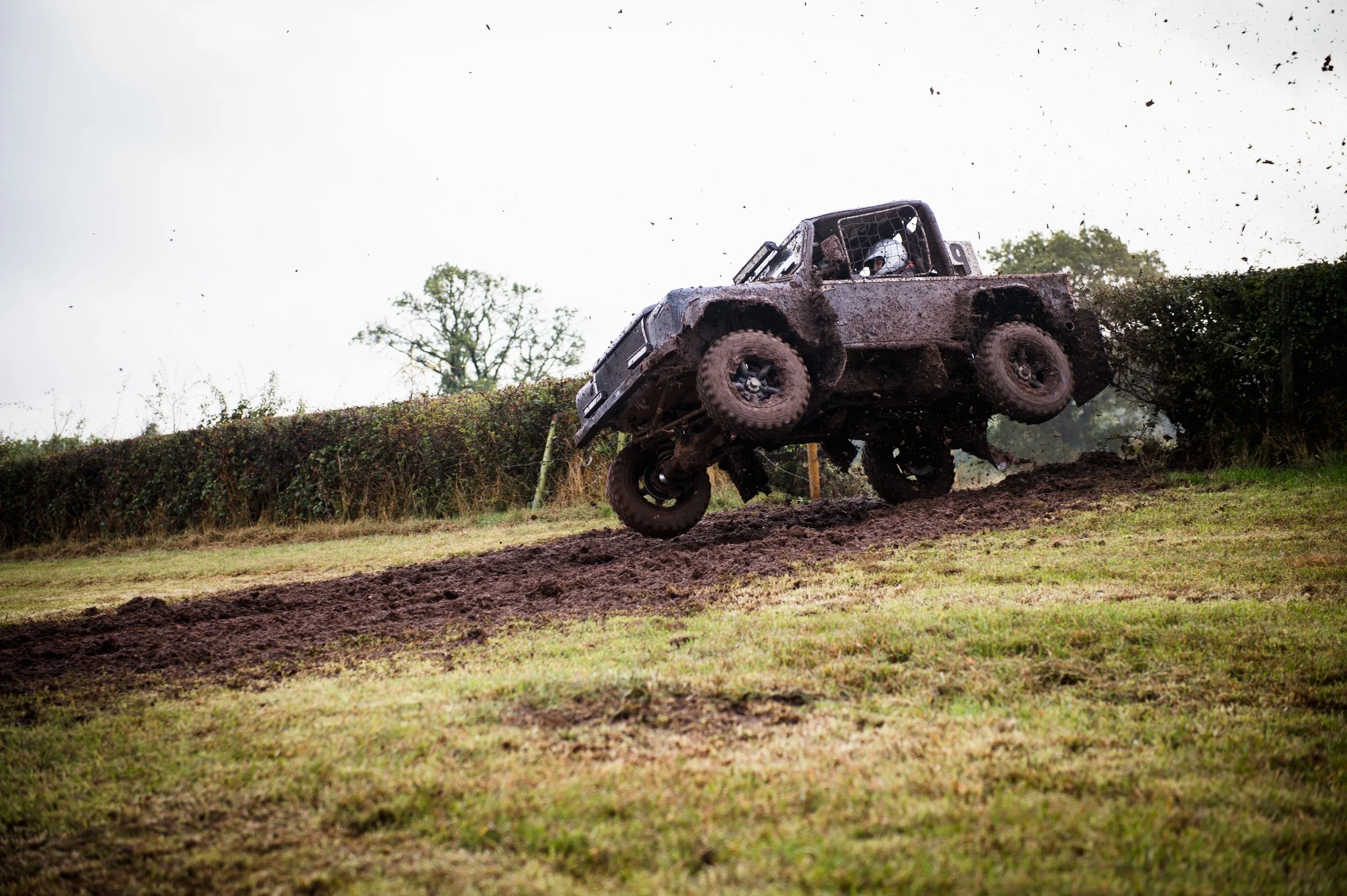 An off-road vehicle with mud splatters jumps over a dirt track during a race, with a driver wearing a helmet.