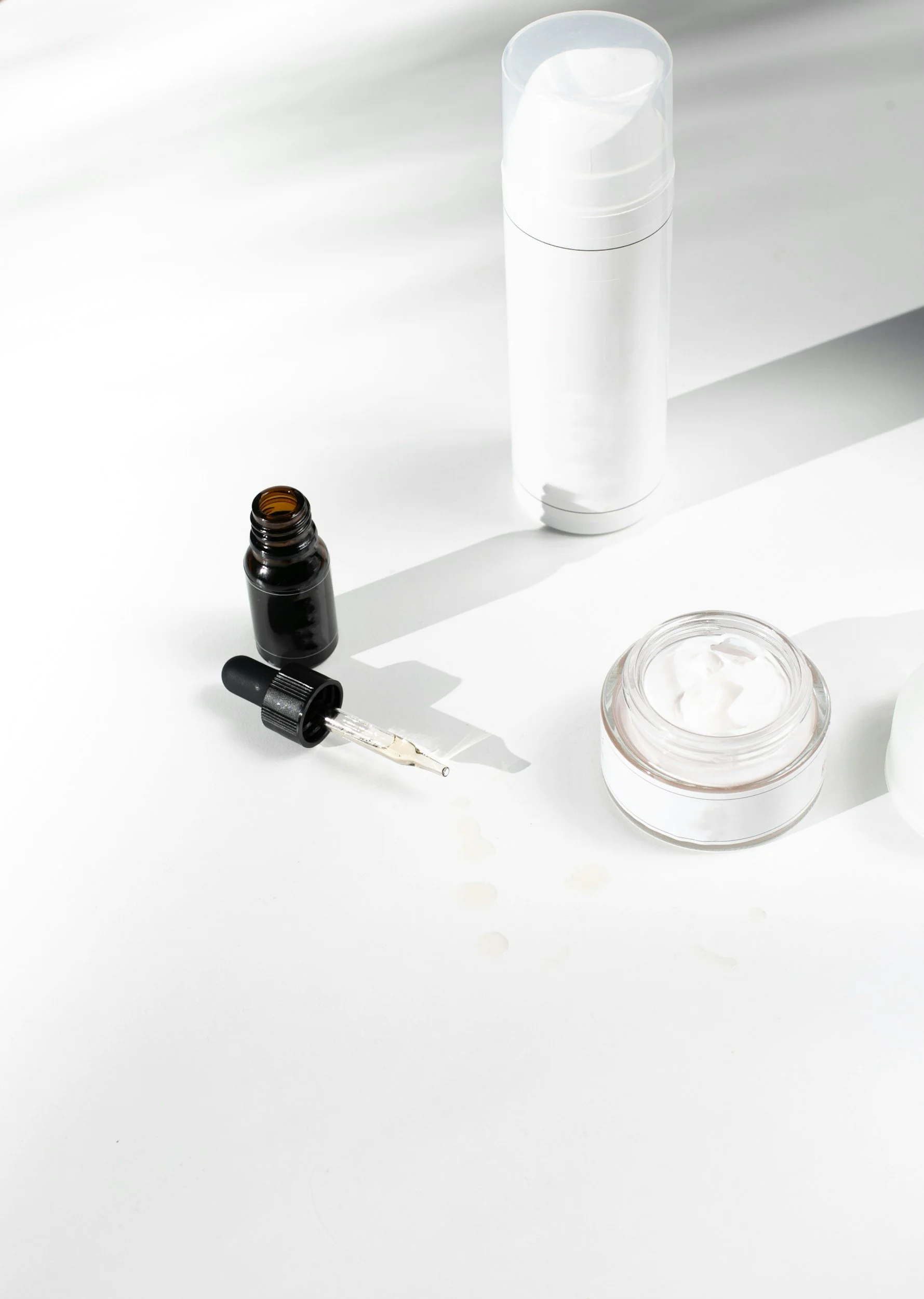 Close-up of skincare products on a white surface, including a small open dark amber bottle with a dropper, a jar of cream, a tall white bottle with a pump, and a small glass container with cream.