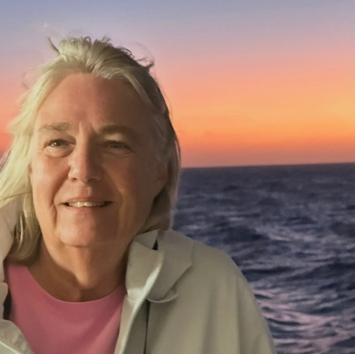 Image of a woman in a white jacket with the ocean and sunset behind her.