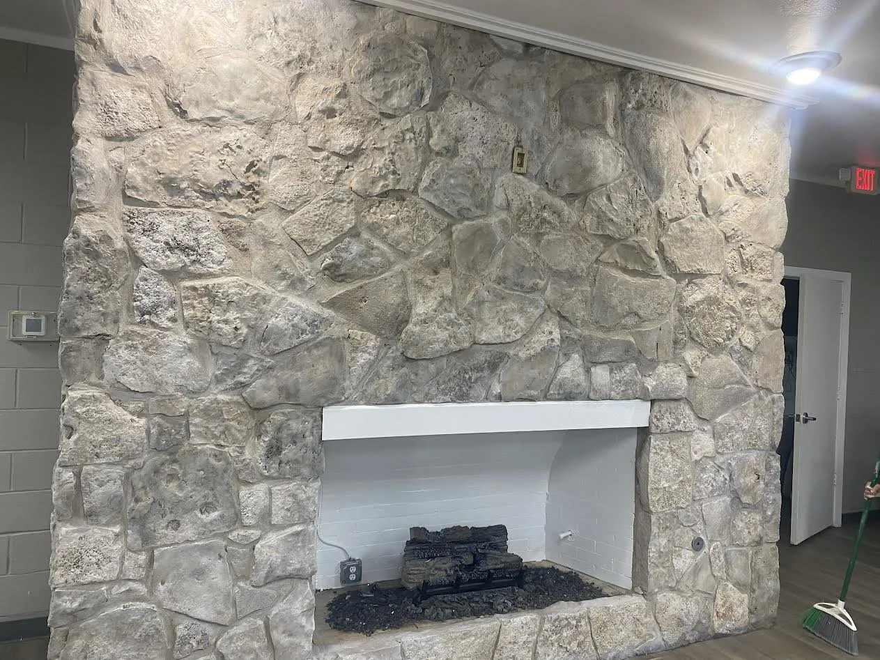 Stone fireplace with white mantel in a room with wood flooring and gray walls, partly visible with a person's hand holding a broom in the corner.