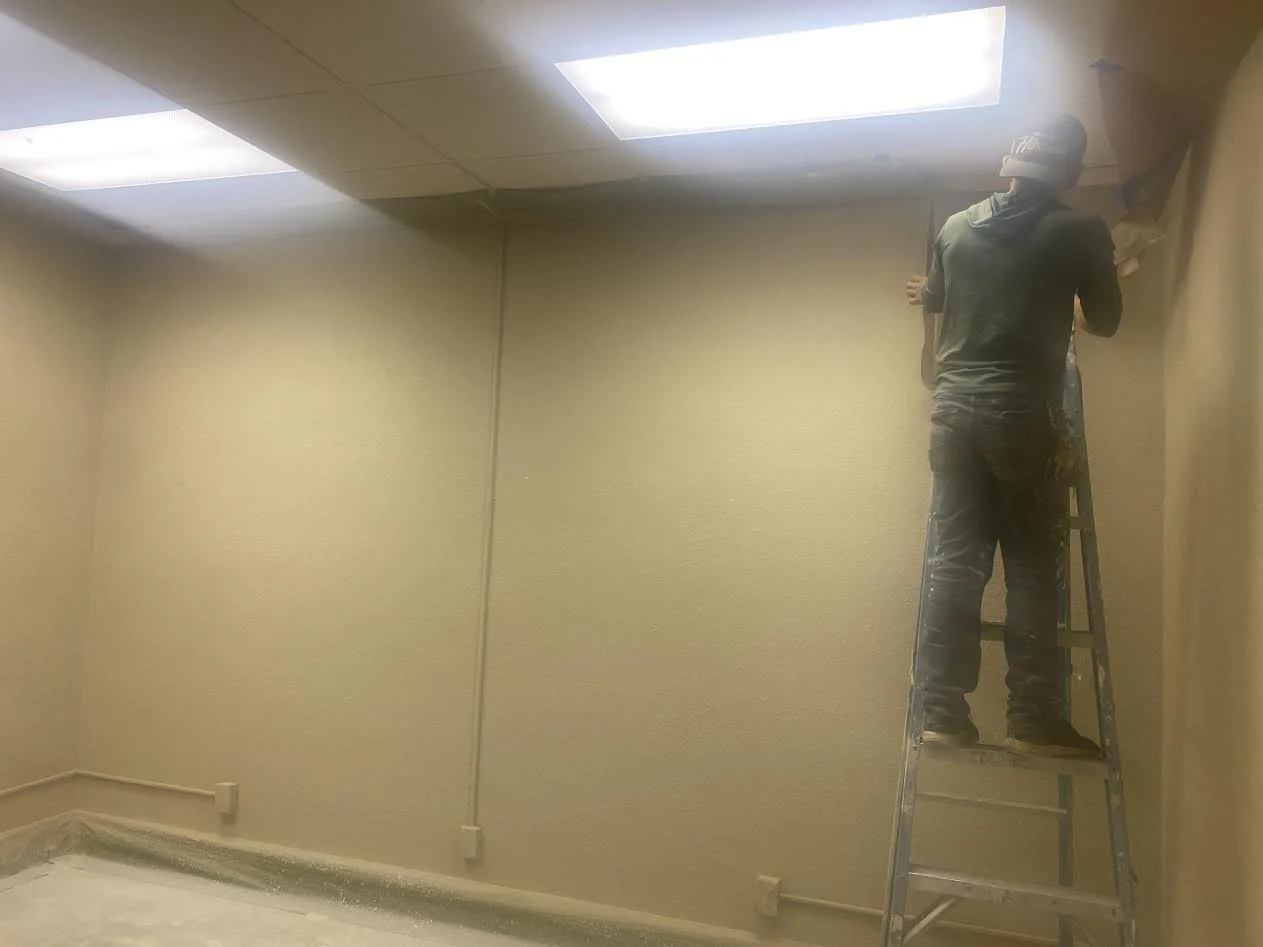 A person standing on a ladder painting a wall in an indoor room with beige walls and fluorescent ceiling lights.