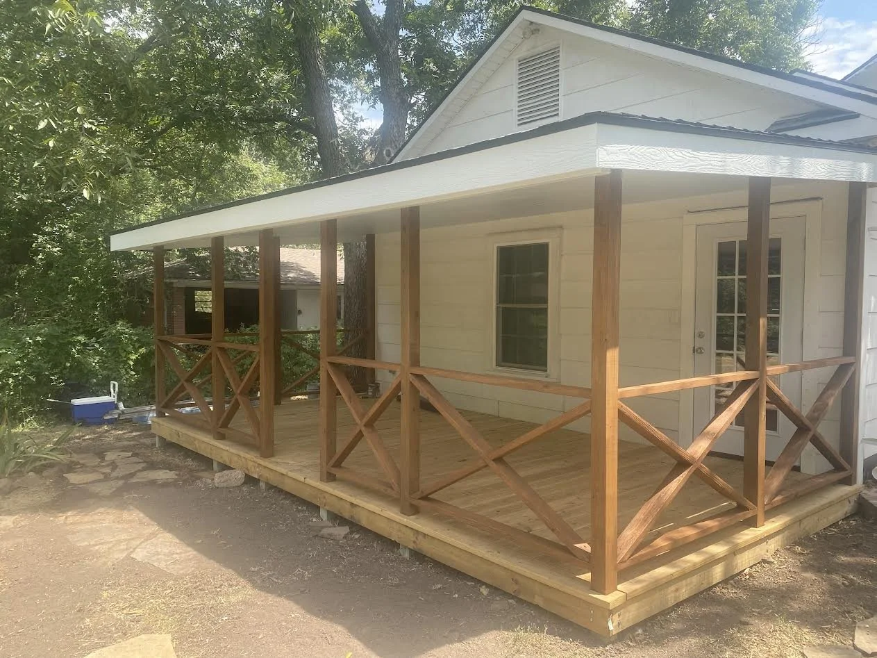 A newly built wooden porch with a railing attached to a white house, surrounded by trees and a dirt pathway.