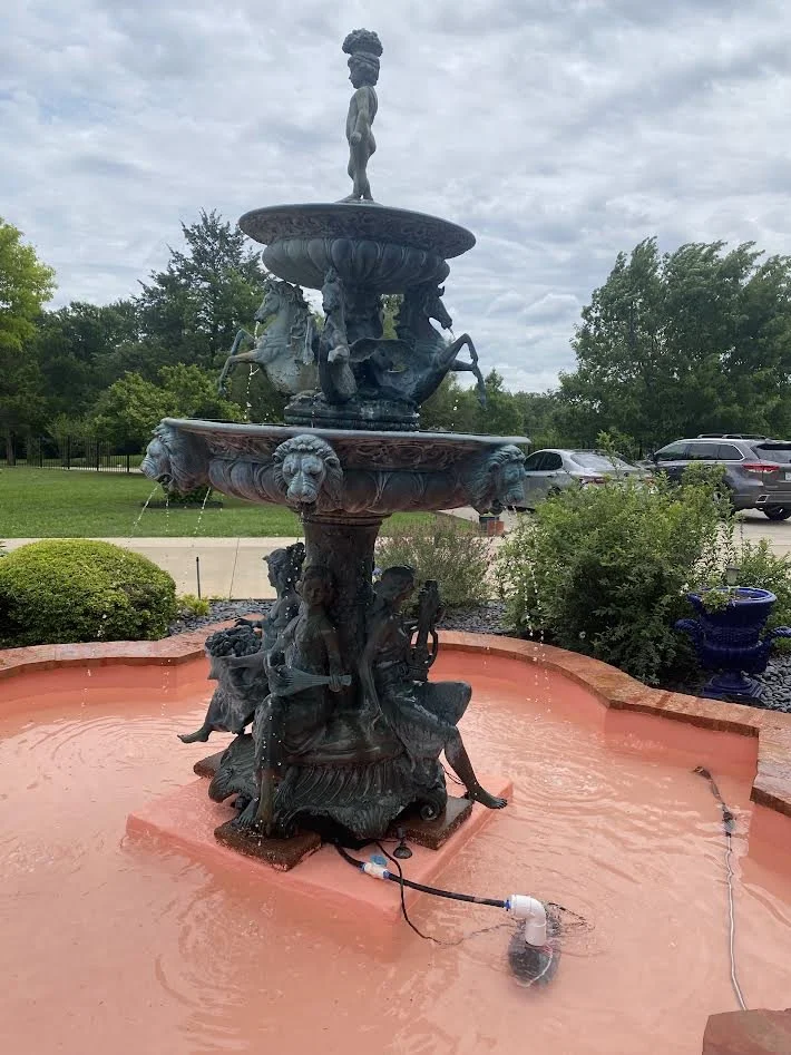 A decorative multi-tiered outdoor fountain with water flowing from sculptures of mermaids, horses, and mythological figures. The fountain has a pinkish-orange basin.