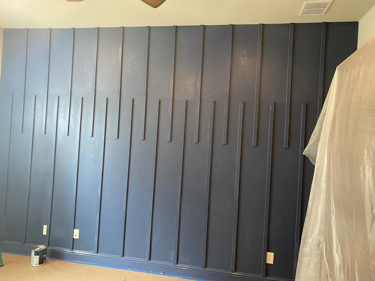 Interior wall with vertical wooden panels painted dark blue, near a bed covered with a beige plastic sheet. A paint can is on the floor at the bottom left corner.
