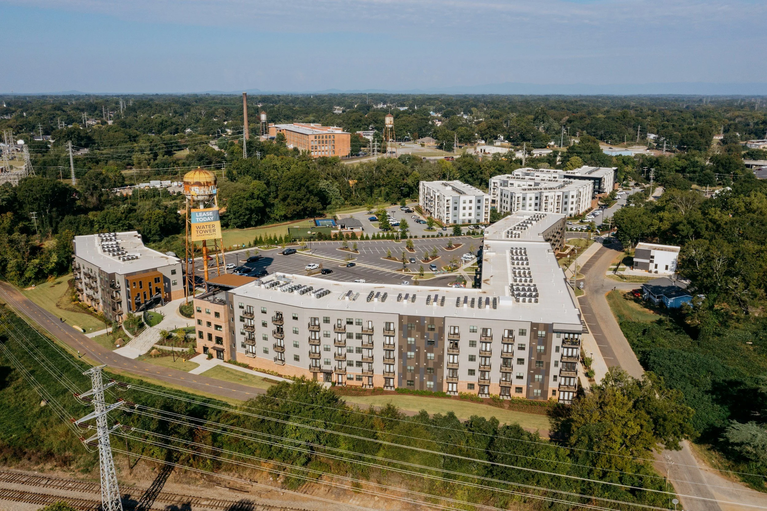 An aerial view of a cityscape featuring modern apartment buildings, a water tower with a sign advertising apartments available for lease, parking lots, trees, and a dense forest in the background. Power lines and electrical towers cross the scene.