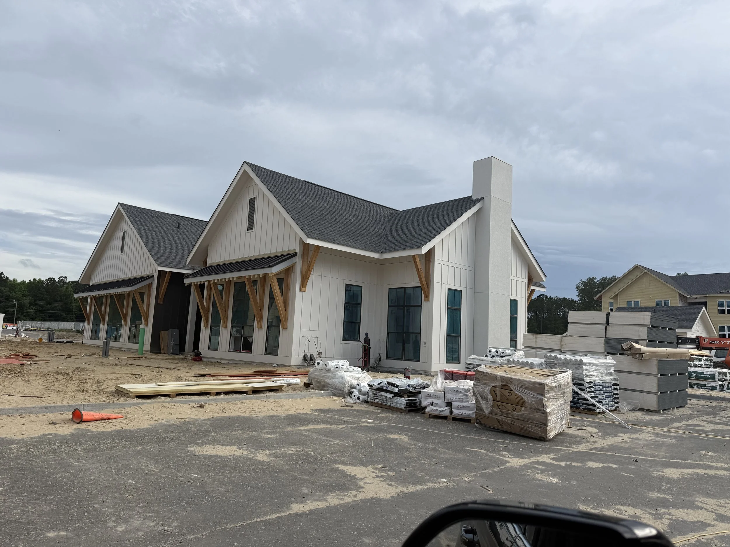 Under construction view of a modern white house with black roof and large windows, surrounded by construction materials and equipment, with a cloudy sky overhead.