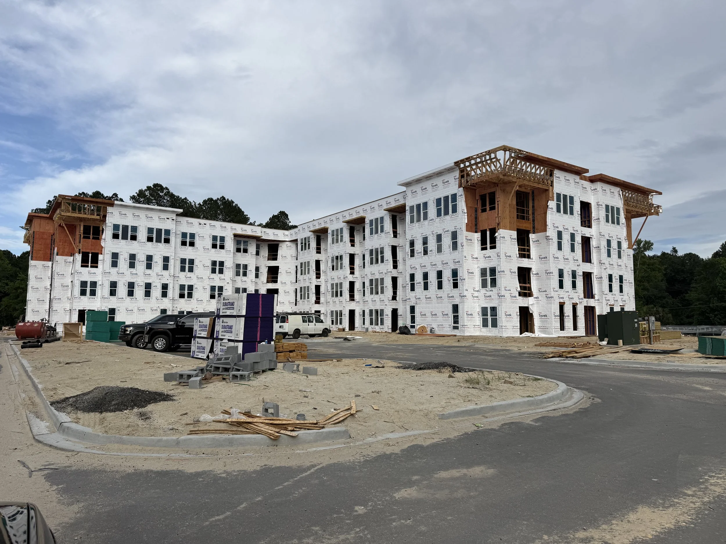 A multi-story residential building under construction with white sheathing and multiple windows, situated in a developing area with construction vehicles, materials, and a partially paved road.