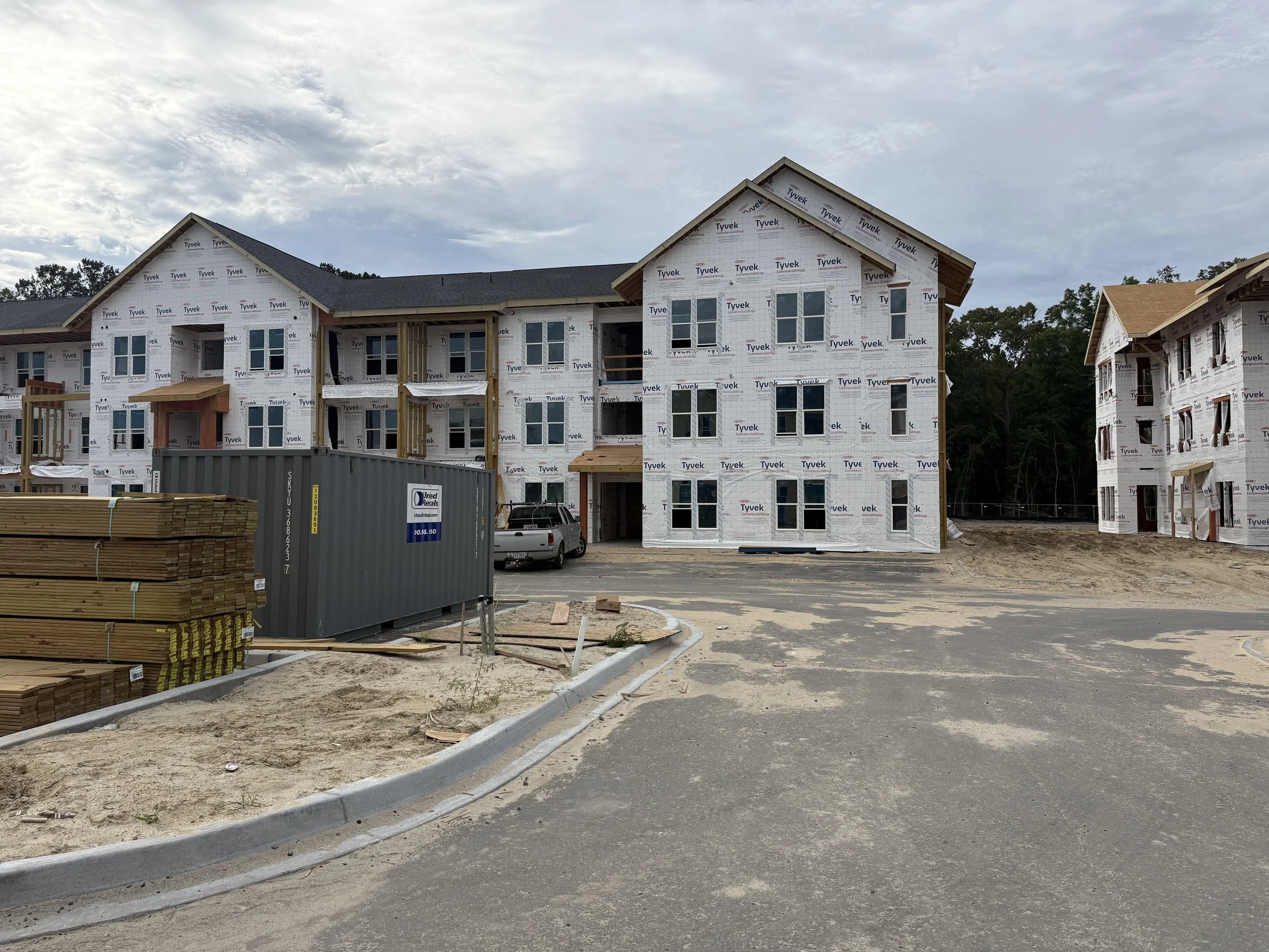 Construction site of residential buildings with incomplete structures covered in white Tyvek weather-resistant wrap, with some windows installed. There are construction materials like lumber, a shipping container, and a vehicle in the foreground. The sky is partly cloudy.