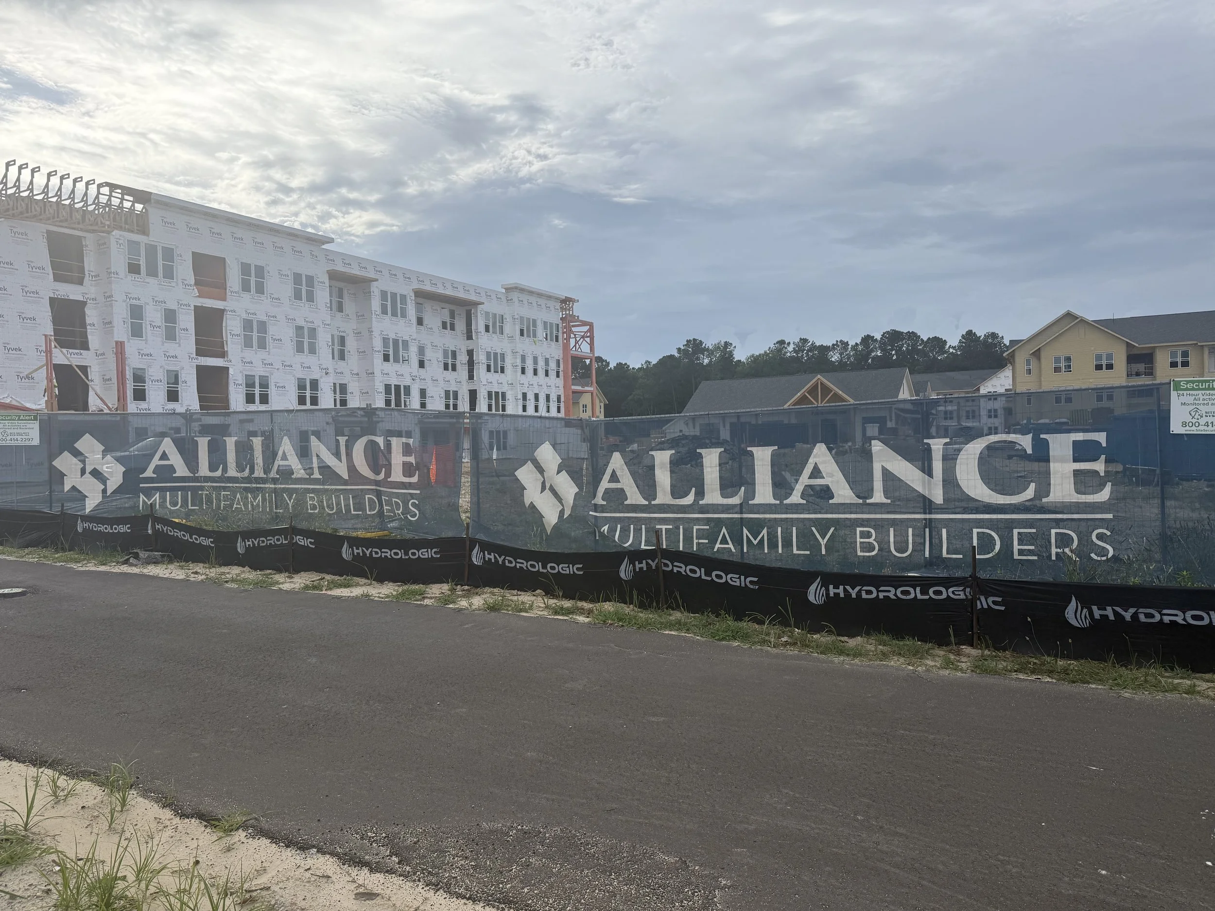 Construction site with partially built multi-story building covered in white sheathing; fencing with signs reading "Alliance Multi-Family Builders" and "Hydrolagic"; neighboring completed yellow house, cloudy sky, and trees in background.