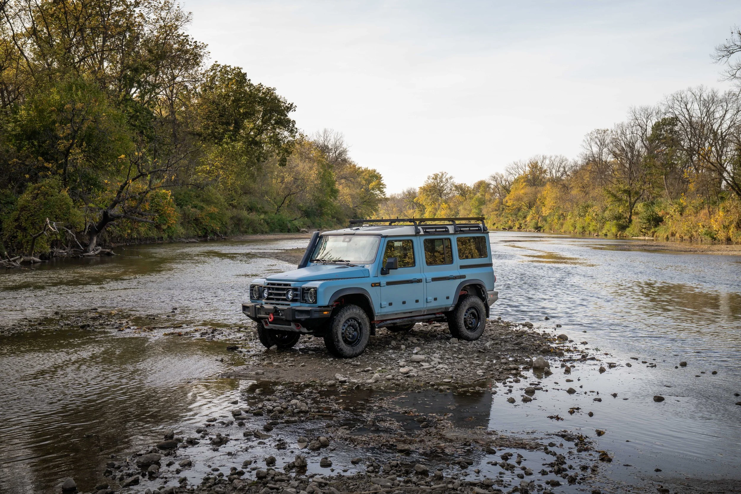 A blue off-road vehicle parked on the rocky edge of a riverbank, with trees with autumn foliage on both sides of the river under a clear sky.