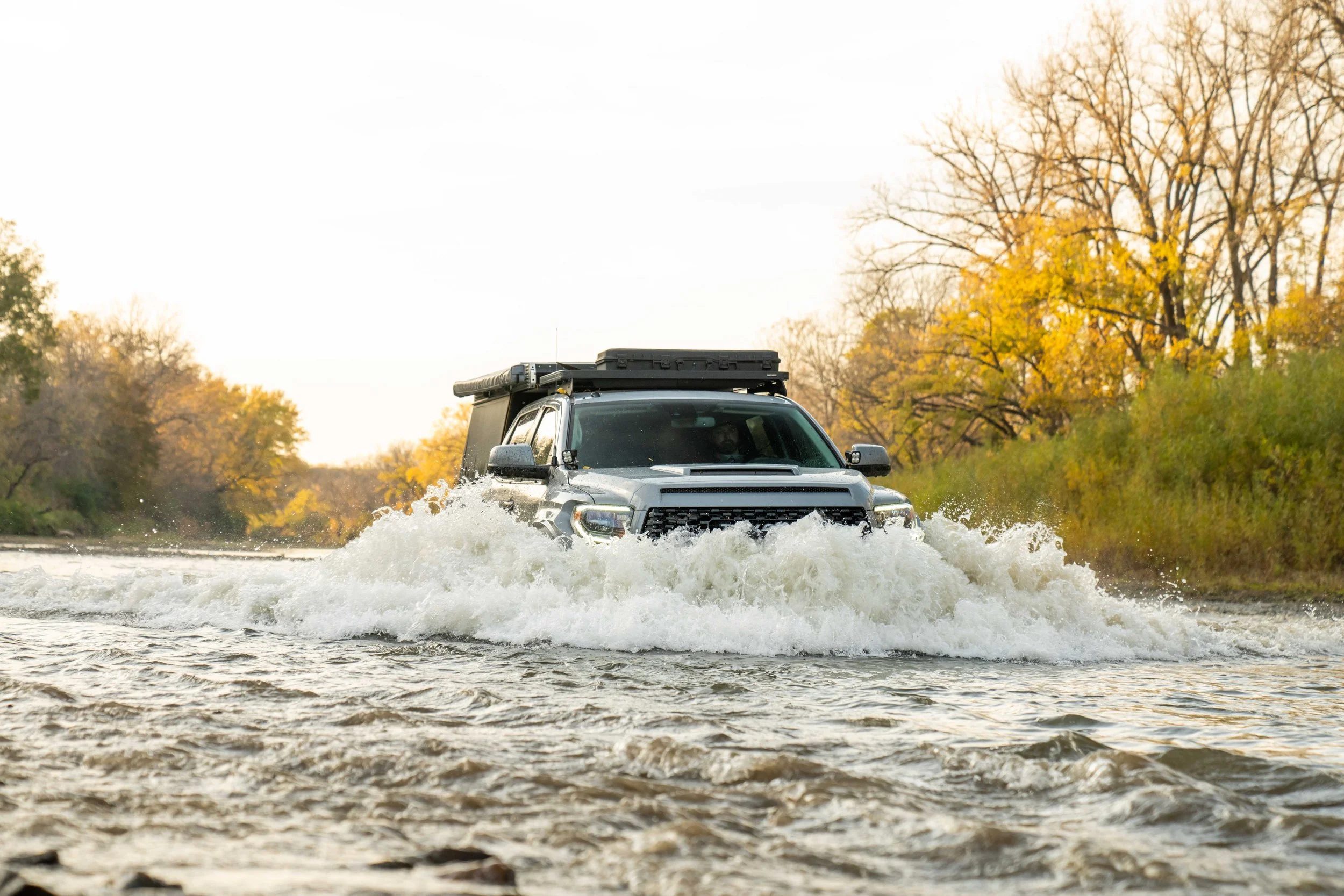 A black SUV driving through a water crossing in a wooded area with fall foliage and trees with yellow and orange leaves.